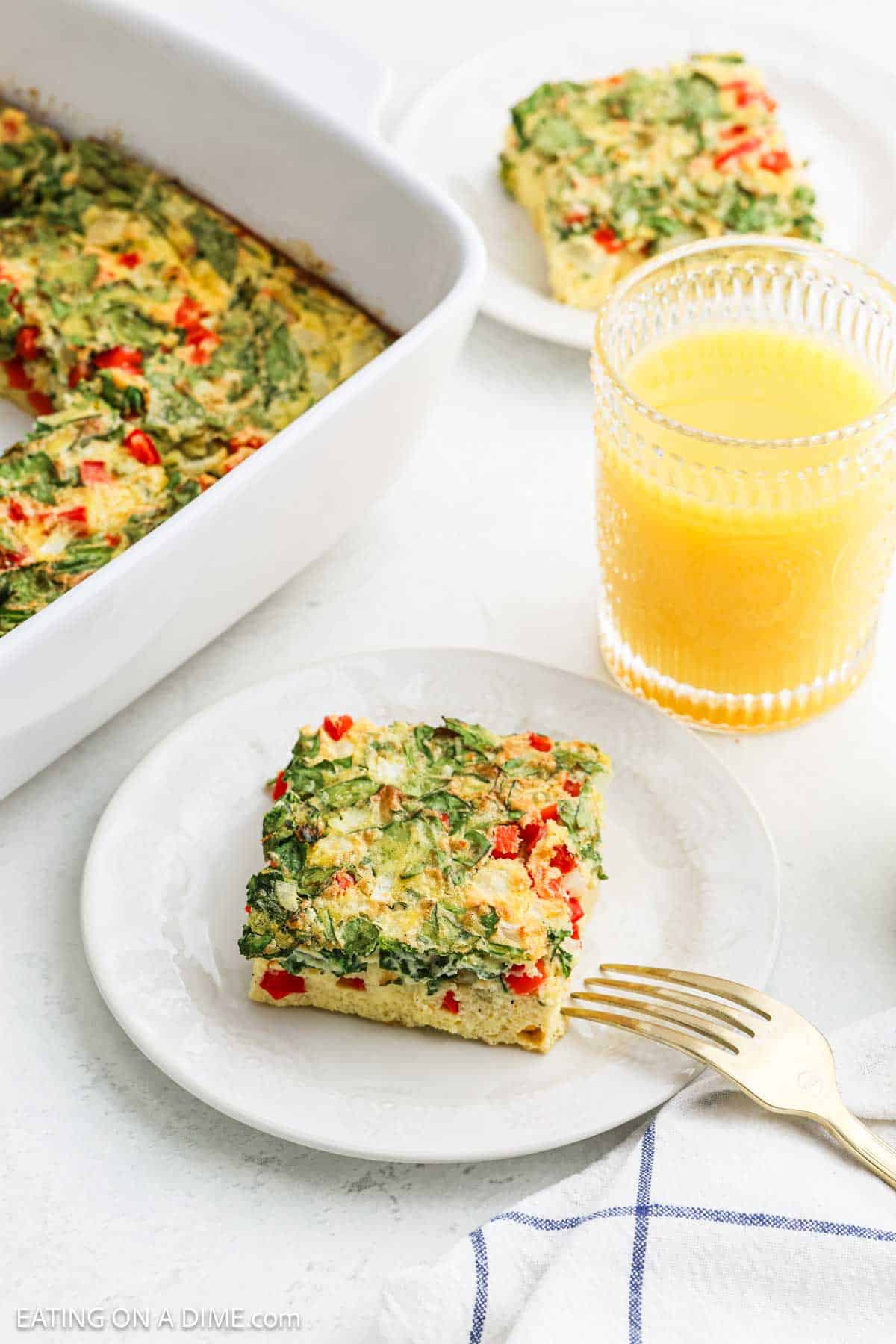 Slice of Egg Casserole with Cottage Cheese on a plate with a fork with a glass of orange juice and egg casserole in a baking dish on the side