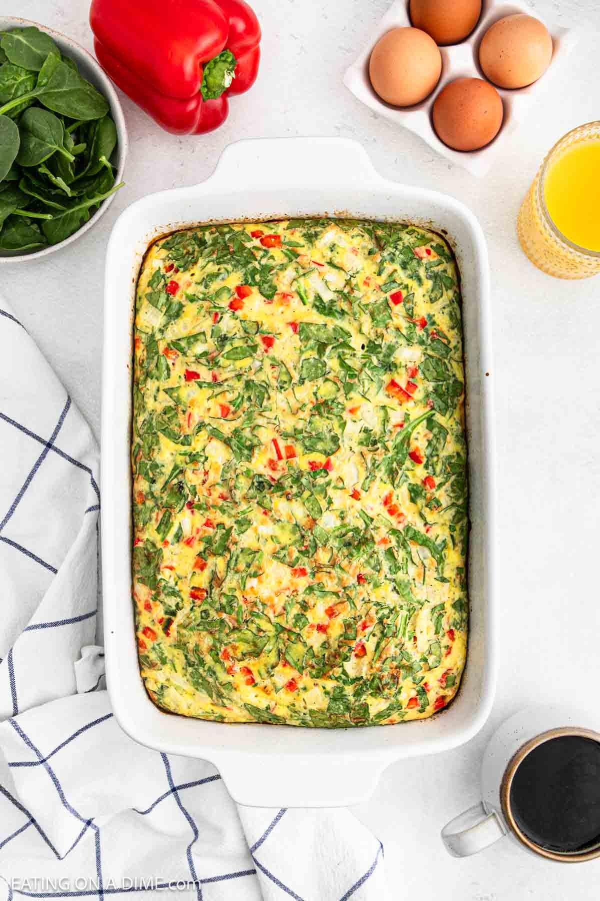 Baked Egg Casserole with Cottage Cheese in a baking dish, with a bowl of spinach, red bell pepper, fresh eggs, orange juice and coffee on the side