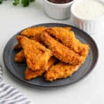 A dark plate filled with crispy, golden-brown oven fried chicken tenders sits on a light surface, with two small bowls of dipping sauces in the background. A striped napkin is partially visible in the corner.