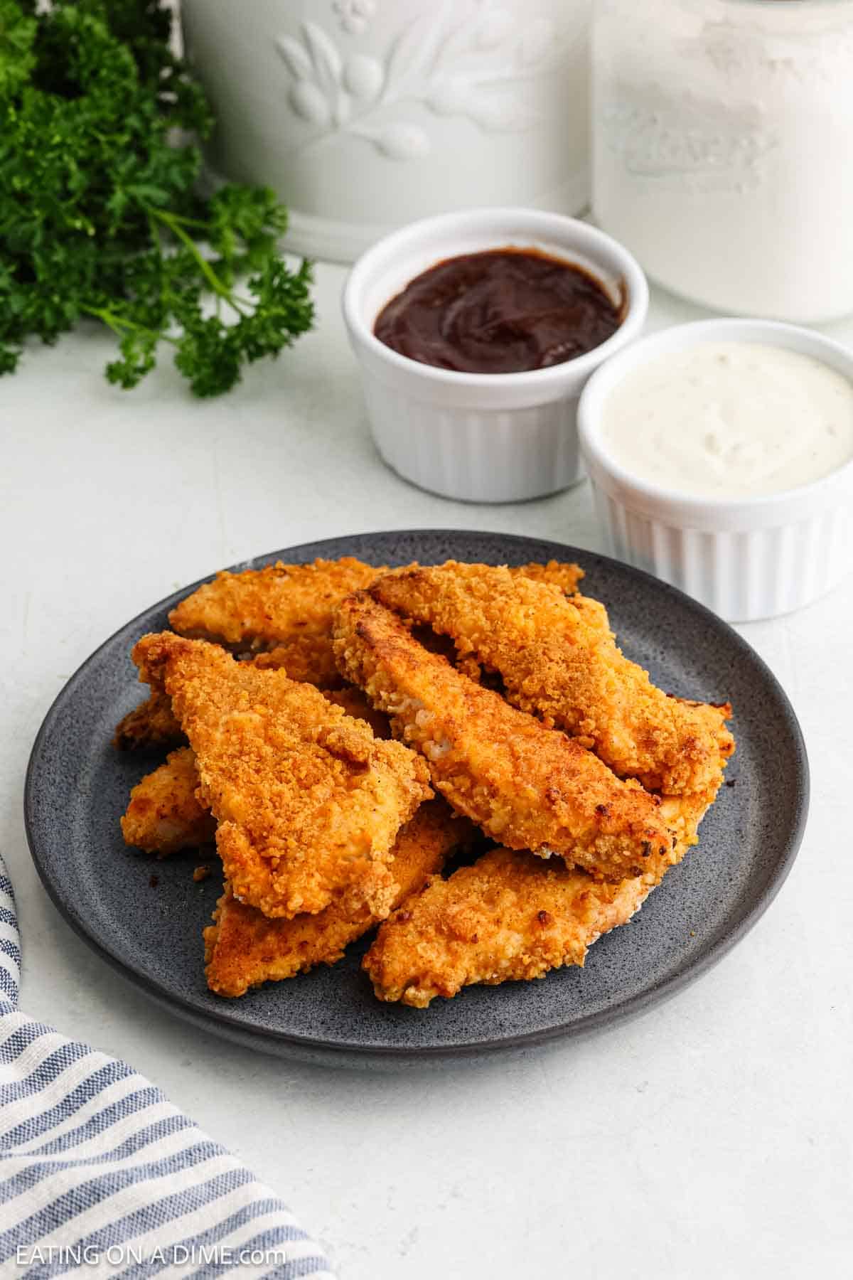 Oven Fried Chicken Tenders stacked on a plate with small bowls of BBQ Sauce and Gravy on the side