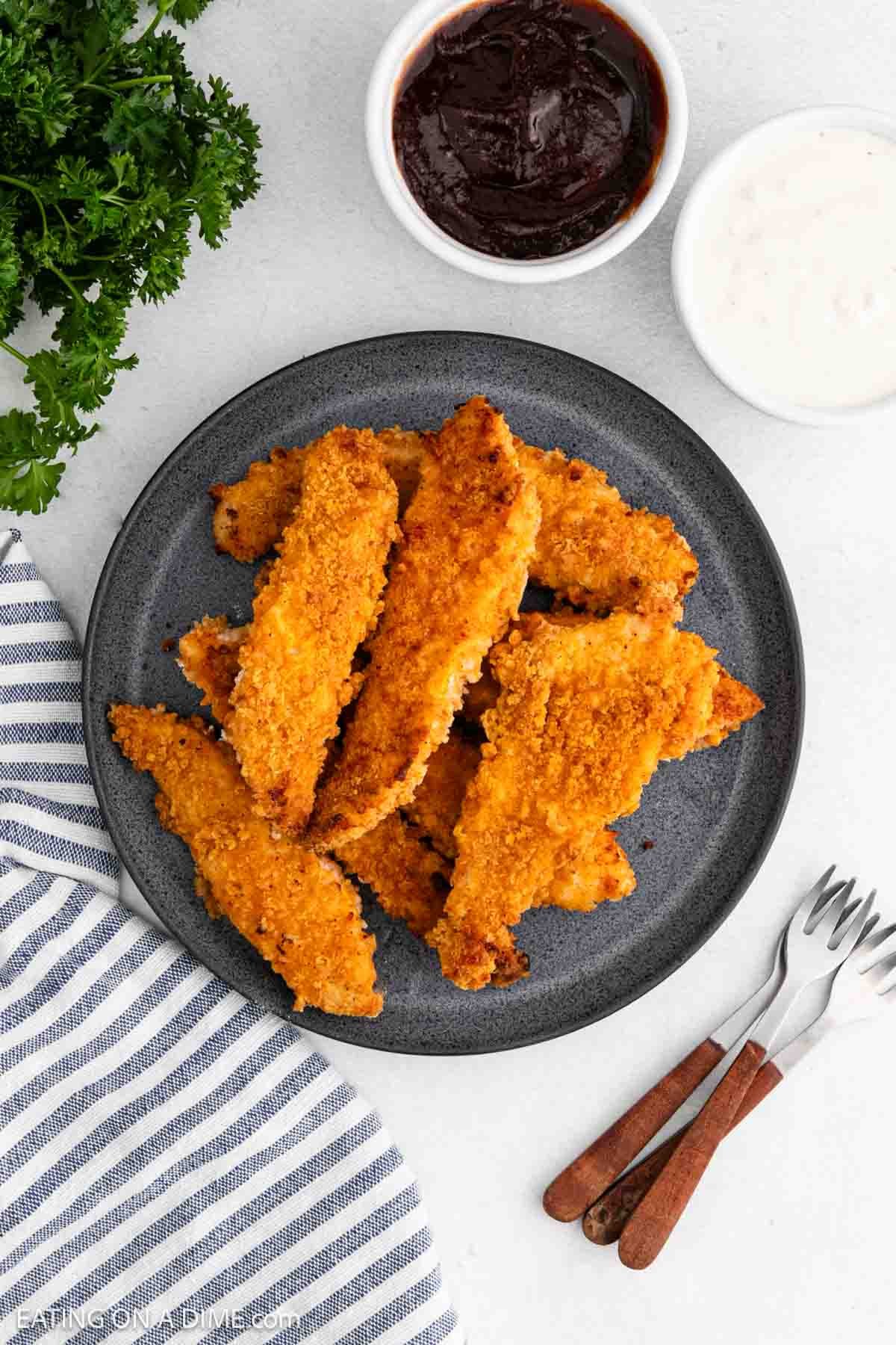 Oven Fried Chicken Tenders stacked on a plate with a bowl of BBQ Sauce and Gravy on the side