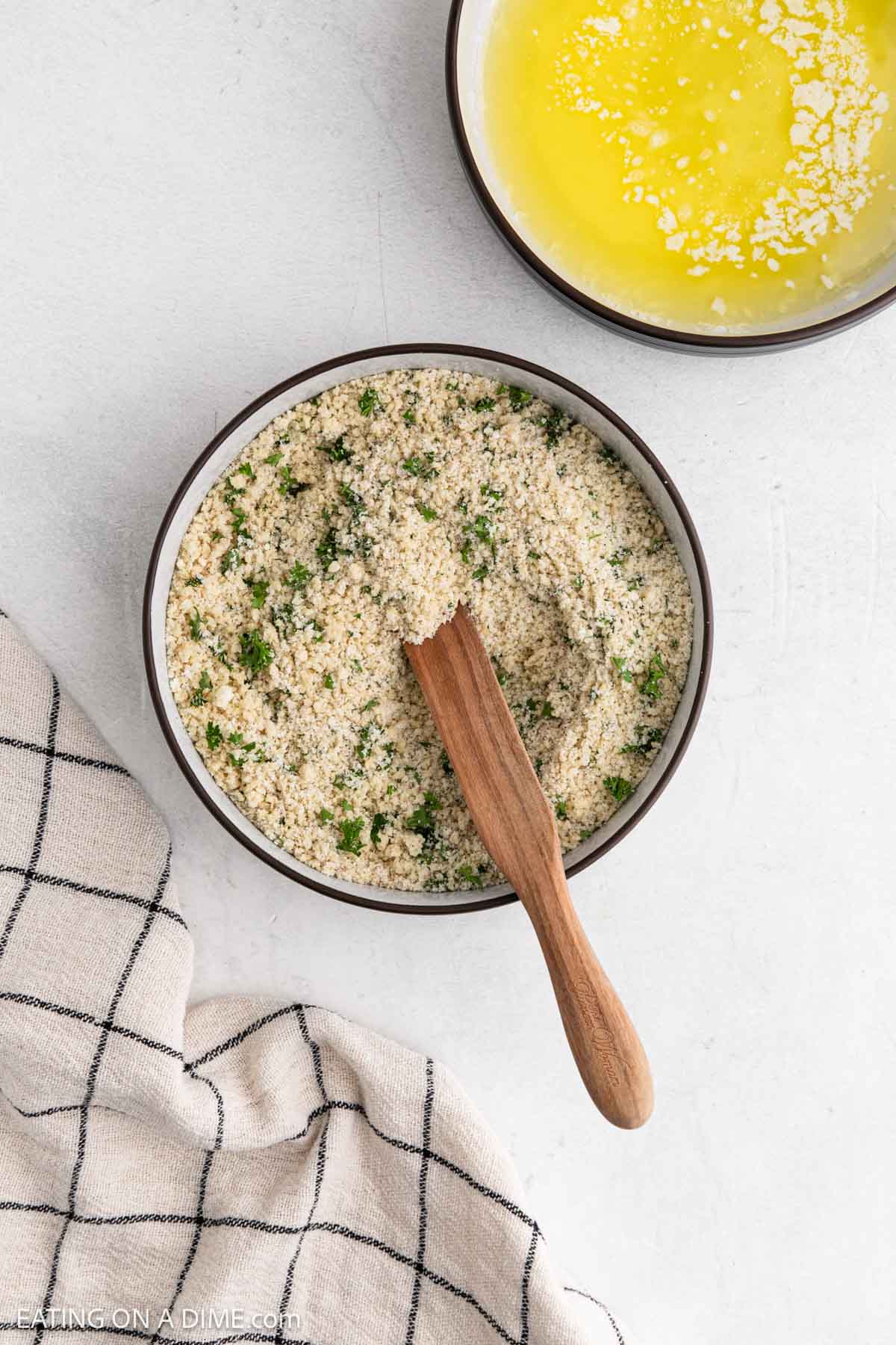 Combining the breadcrumbs, parmesan cheese, parsley, salt, pepper, and garlic powder together in a bowl with a wooden spoon with a bowl of melted butter on the side
