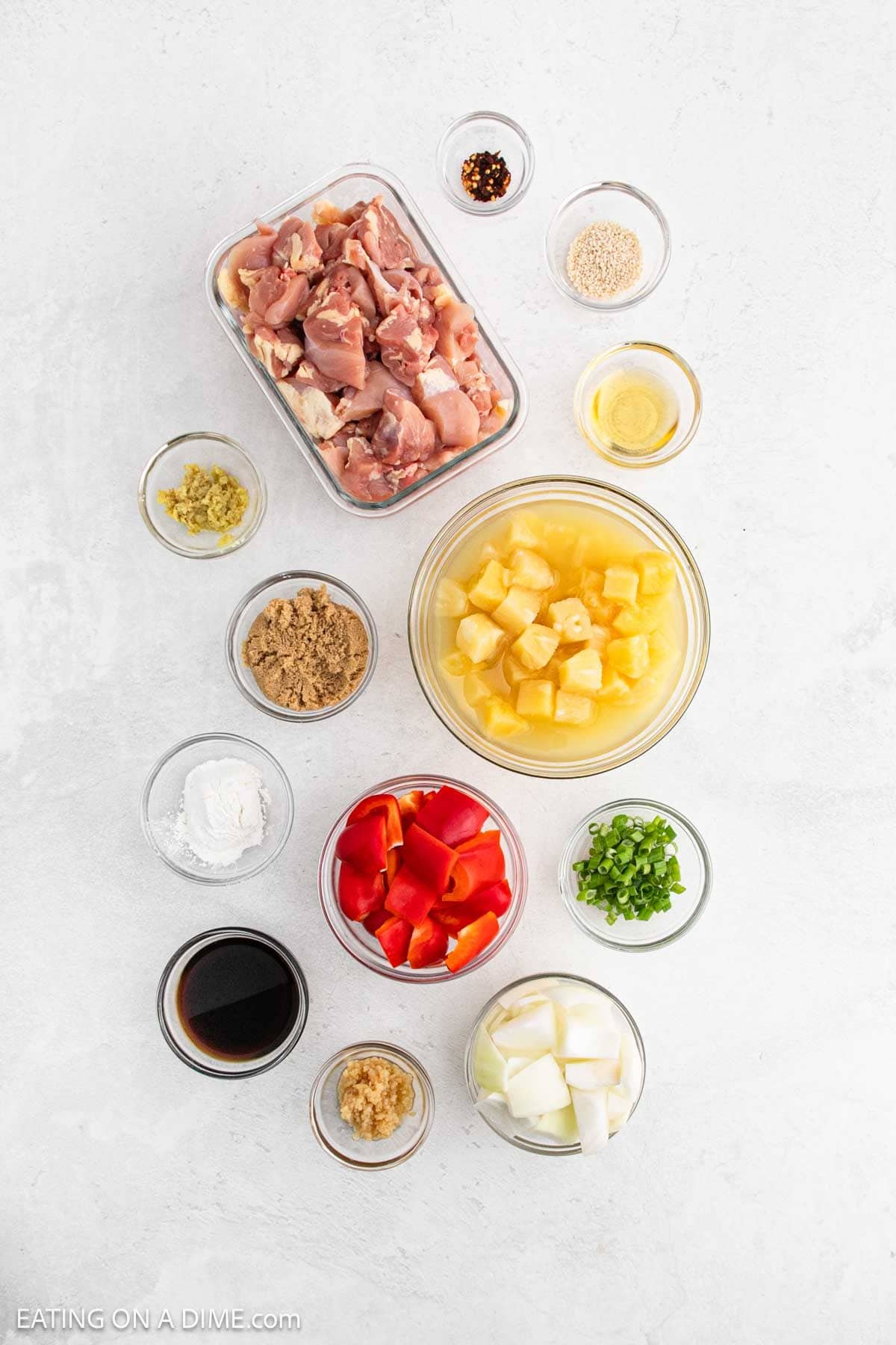 Top-down view of ingredients in bowls for Slow Cooker Pineapple Chicken, featuring raw chicken, pineapple chunks, bell pepper, onion, green onions, brown sugar, soy sauce, ginger, garlic, cornstarch, sesame seeds, red pepper flakes, and vinegar.