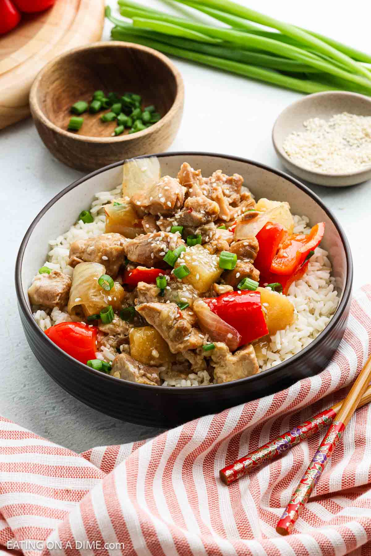 A bowl of white rice topped with Slow Cooker Pineapple Chicken, red bell peppers, pineapple, and green onions, served with chopsticks. Green onions, a bowl of chopped scallions, and sesame seeds are in the background.