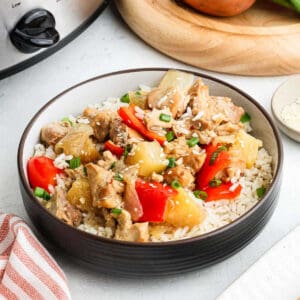 A bowl of white rice topped with Pineapple Chicken—tender chunks of chicken, pineapple, red bell peppers, and green onions—with a slow cooker and a wooden cutting board in the background.