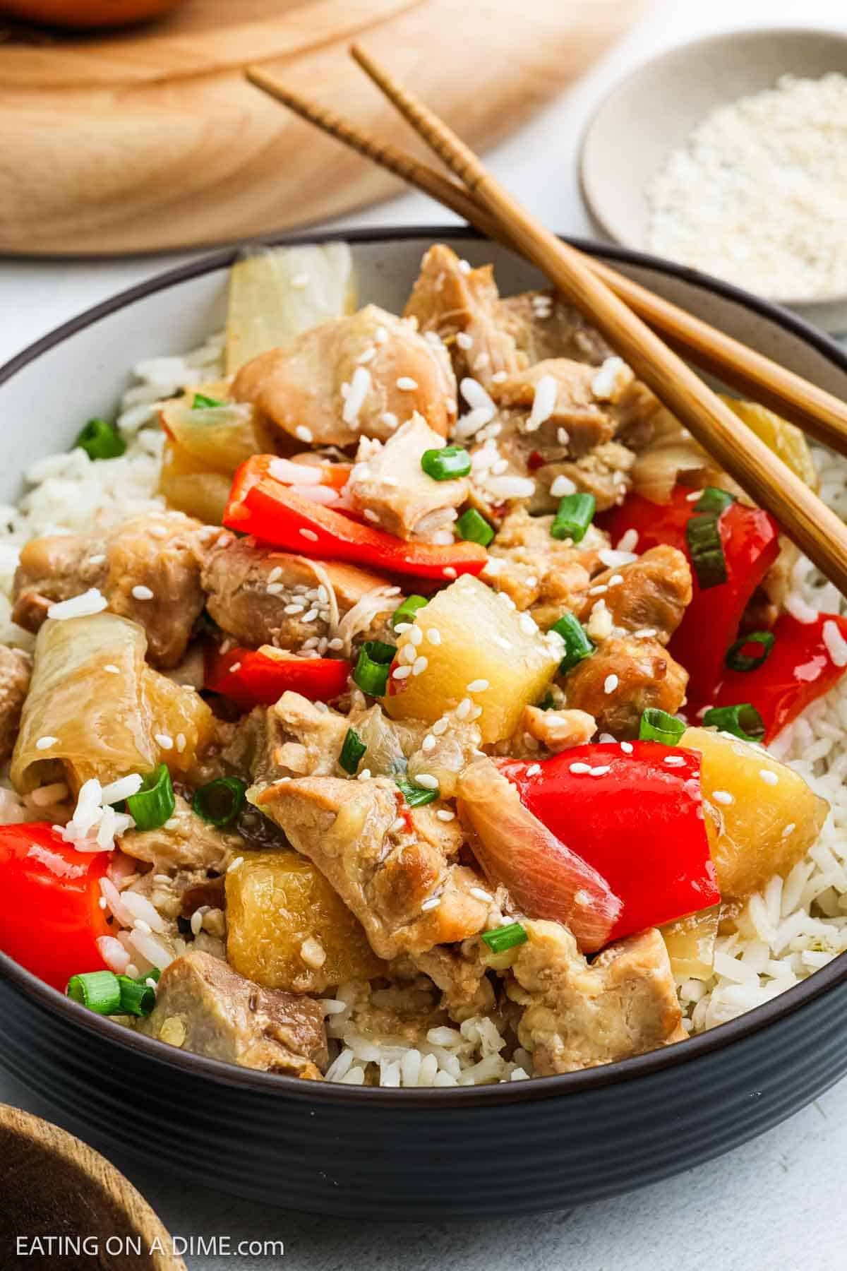 A bowl of slow cooker pineapple chicken served over white rice, topped with red bell peppers, pineapple chunks, green onions, and sesame seeds, accompanied by chopsticks.