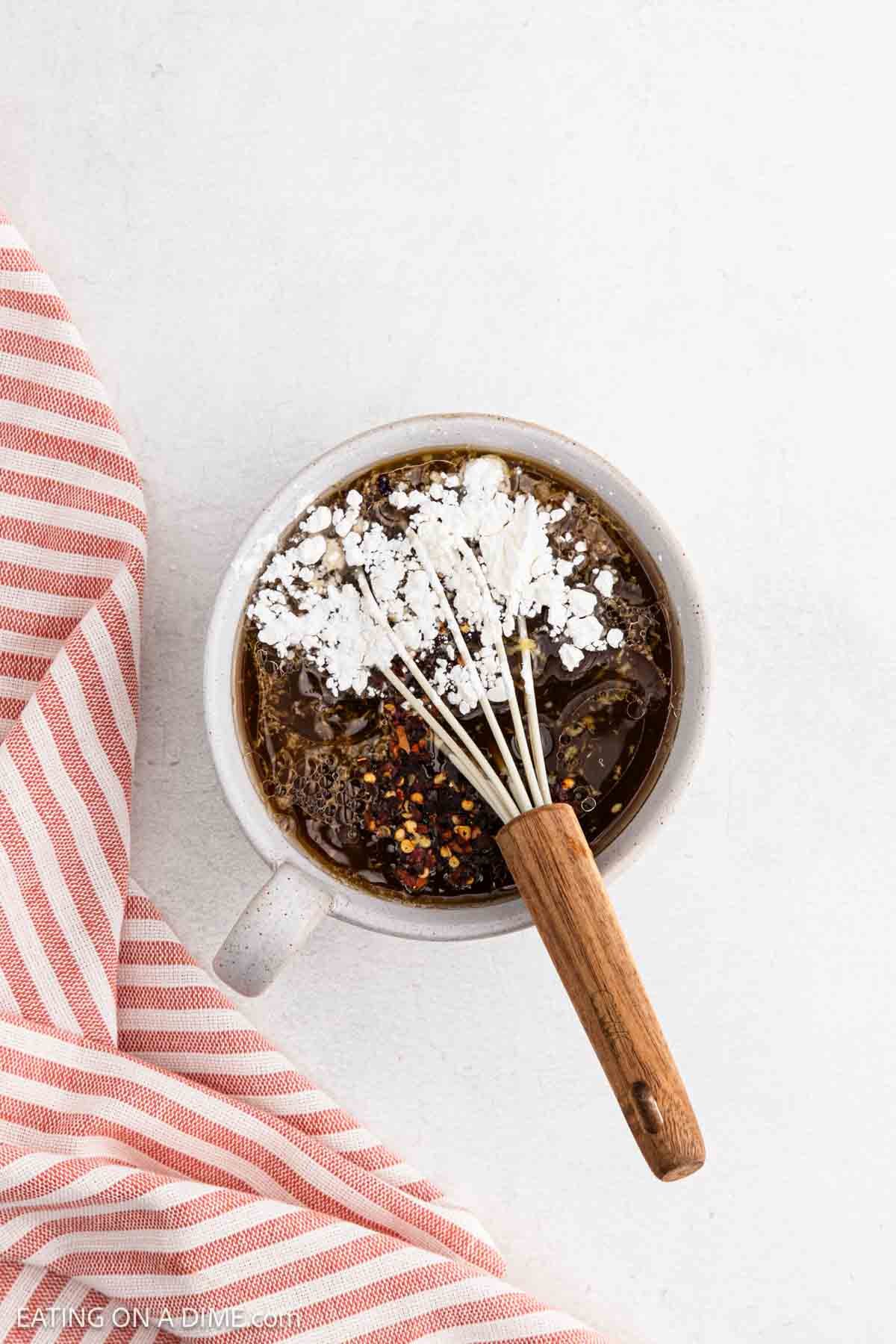 A small white bowl filled with dark liquid, topped with white powder and red pepper flakes, sits beside a red and white striped cloth—a whisk with a wooden handle rests inside, perfect for prepping Slow Cooker Chicken marinades on a clean white surface.