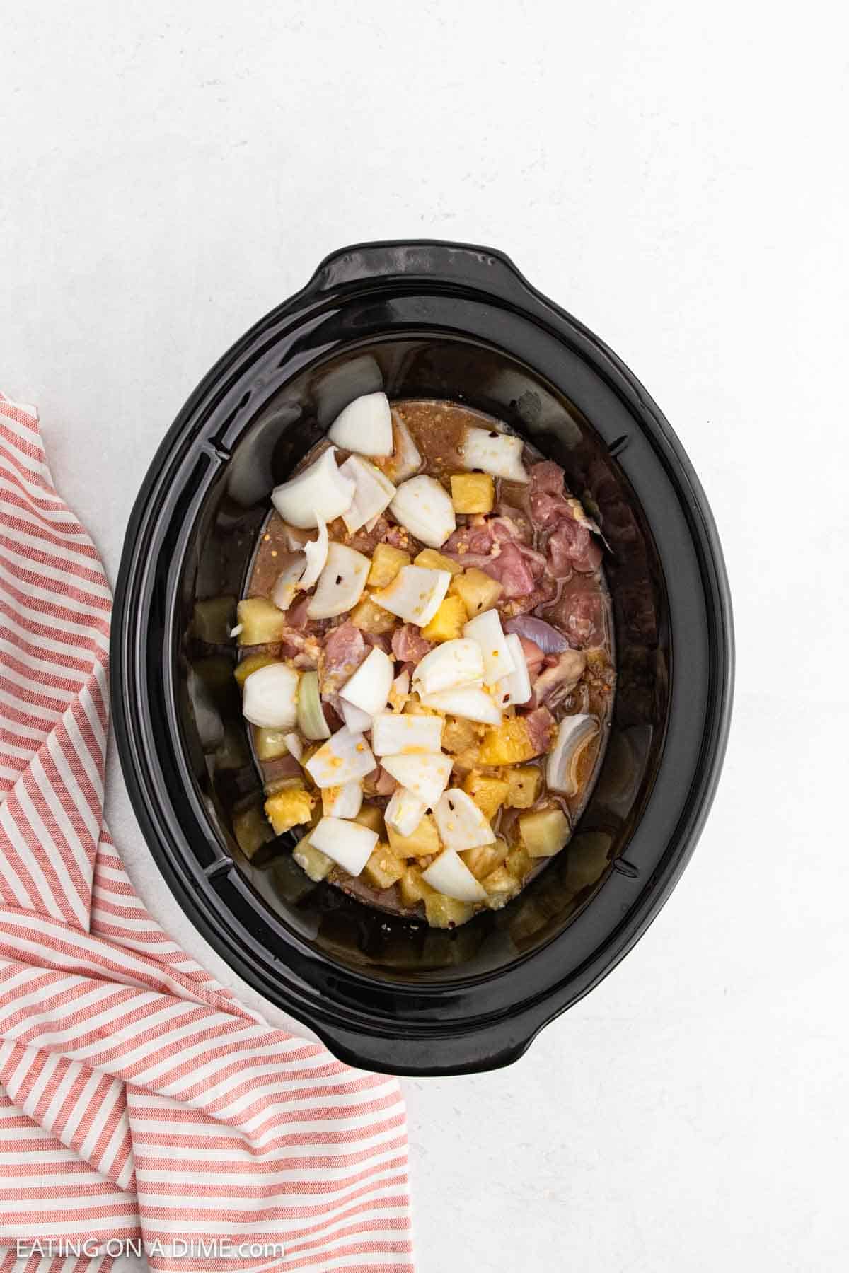 A slow cooker filled with chunks of Slow Cooker Chicken, diced onions, and pineapple pieces sits beside a pink and white striped cloth on a white surface.