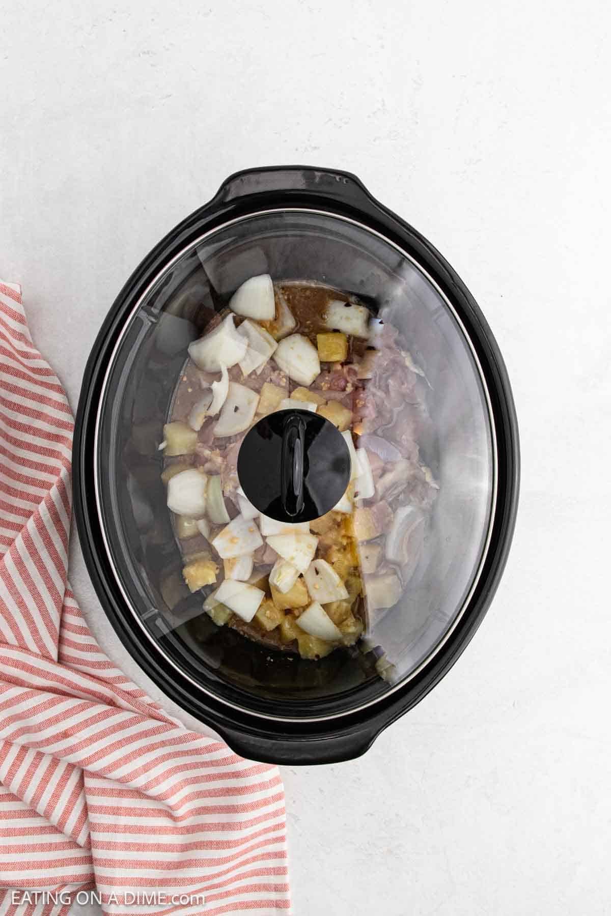 A slow cooker filled with chopped onions, potatoes, and Slow Cooker Chicken, seen from above, sits on a white surface next to a pink and white striped cloth.
