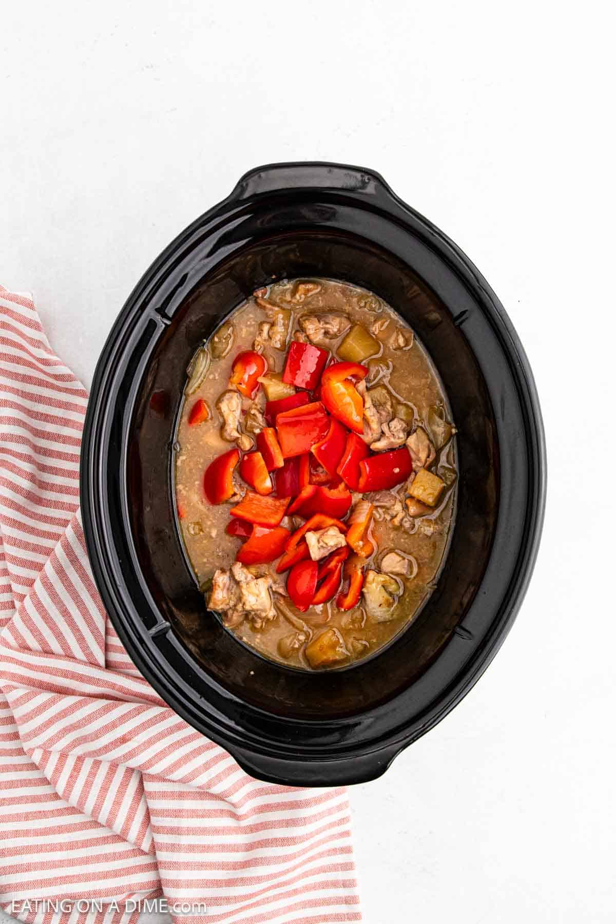 A slow cooker filled with a savory pineapple chicken stew, rich sauce, and chopped red bell peppers sits on a white surface next to a pink and white striped cloth.