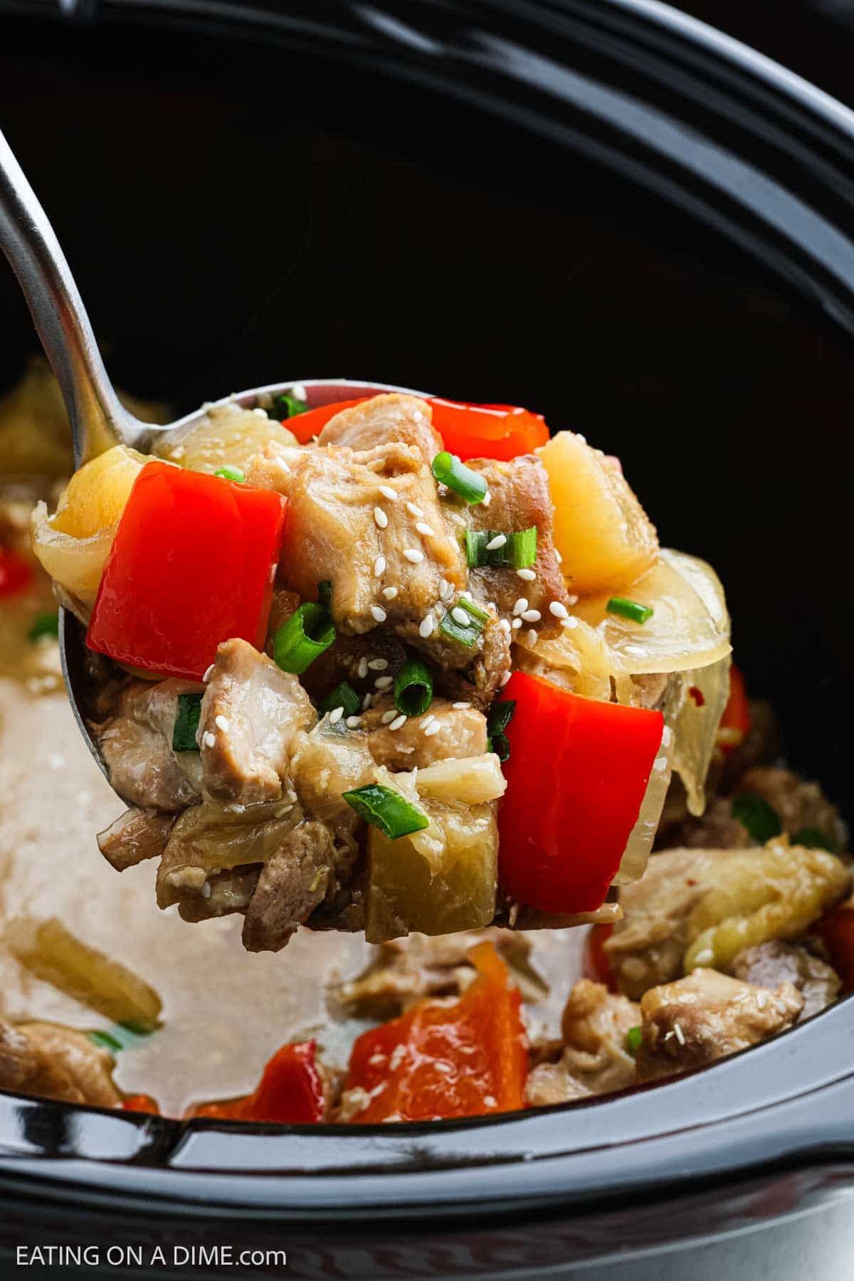 A close-up of a spoonful of slow cooker Pineapple Chicken with red bell peppers, pineapple, onions, green onions, and sesame seeds, held above a slow cooker filled with more of the dish.
