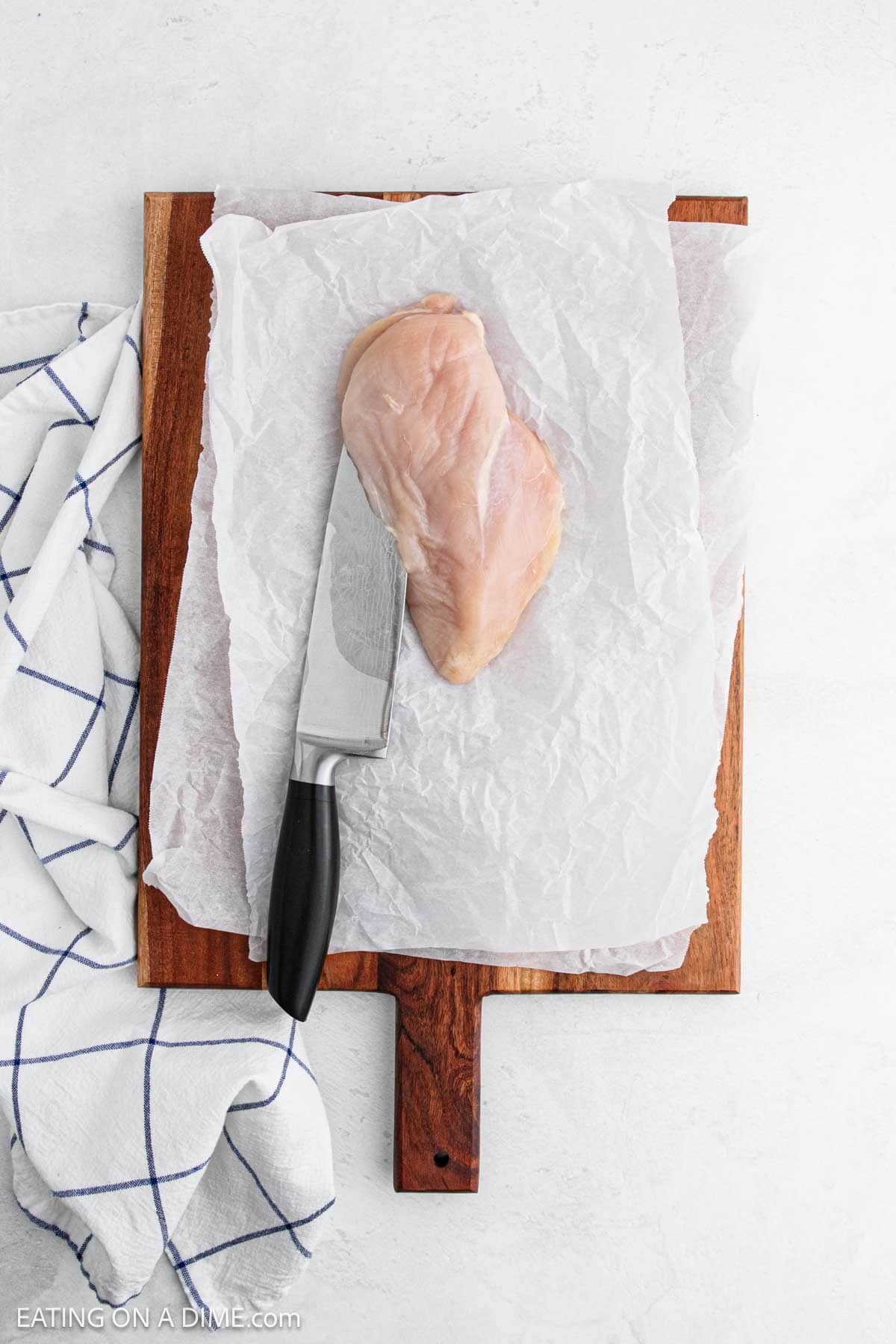 A raw spinach stuffed chicken breast sits on parchment paper atop a wooden cutting board, with a chef’s knife beside it. A white kitchen towel with blue stripes is partially visible on the left.