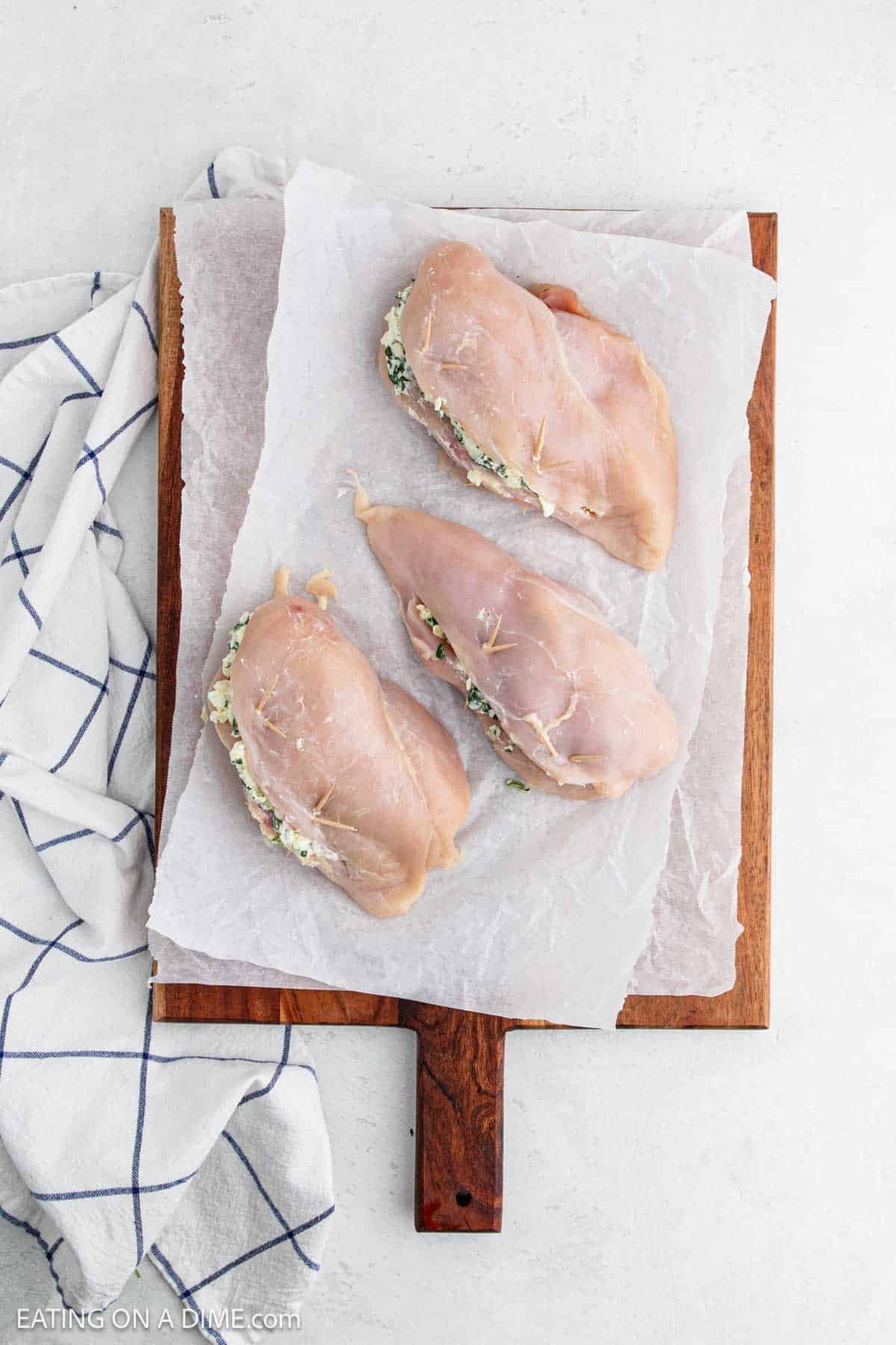 Three raw spinach stuffed chicken breasts with a creamy filling rest on parchment paper atop a wooden cutting board, next to a white kitchen towel with blue stripes.