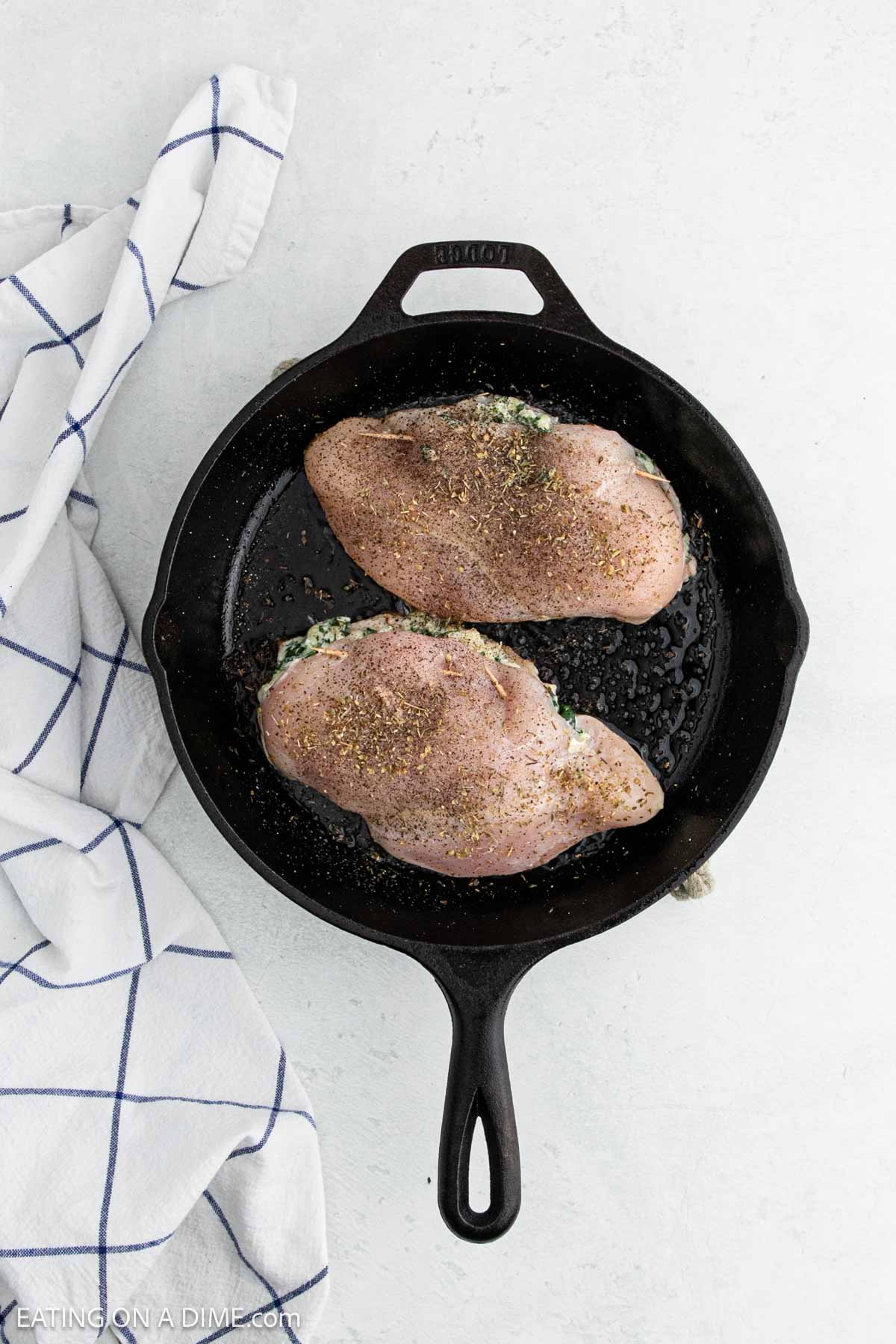 A black cast iron skillet holds two seasoned raw chicken breasts, partially stuffed with a green herb mixture for a delicious Spinach Stuffed Chicken Breast. A white kitchen towel with blue stripes rests nearby on a light surface.
