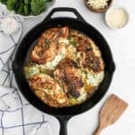 Four cooked, golden-brown Spinach Stuffed Chicken Breasts filled with creamy spinach and cheese in a black skillet, surrounded by fresh spinach leaves, a bowl of shredded cheese, garlic, and a wooden spatula on a white surface.