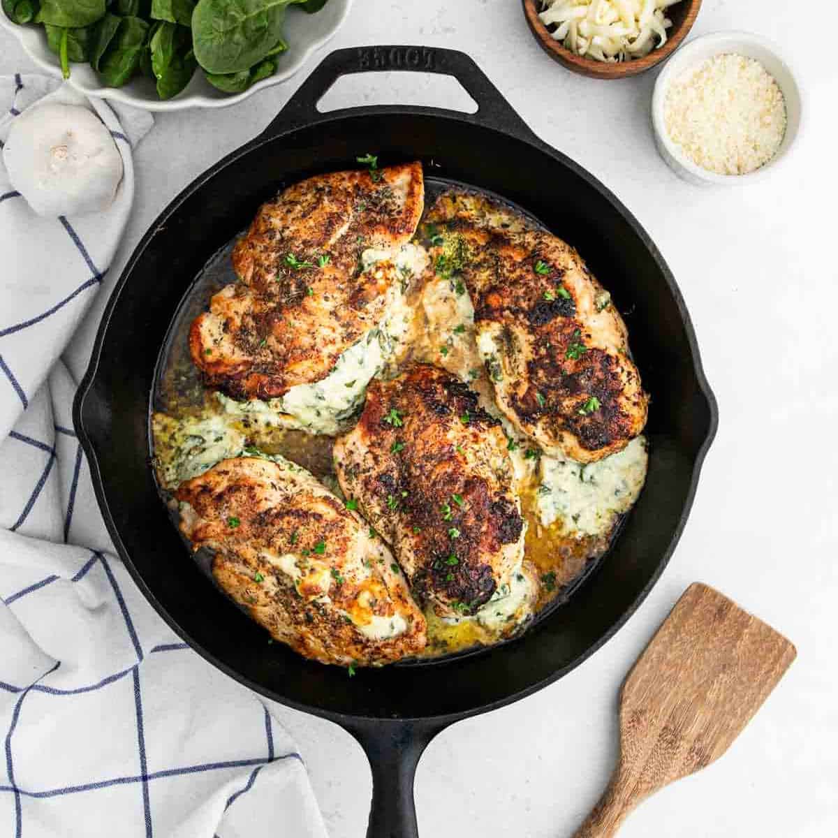 Four cooked, golden-brown Spinach Stuffed Chicken Breasts filled with creamy spinach and cheese in a black skillet, surrounded by fresh spinach leaves, a bowl of shredded cheese, garlic, and a wooden spatula on a white surface.