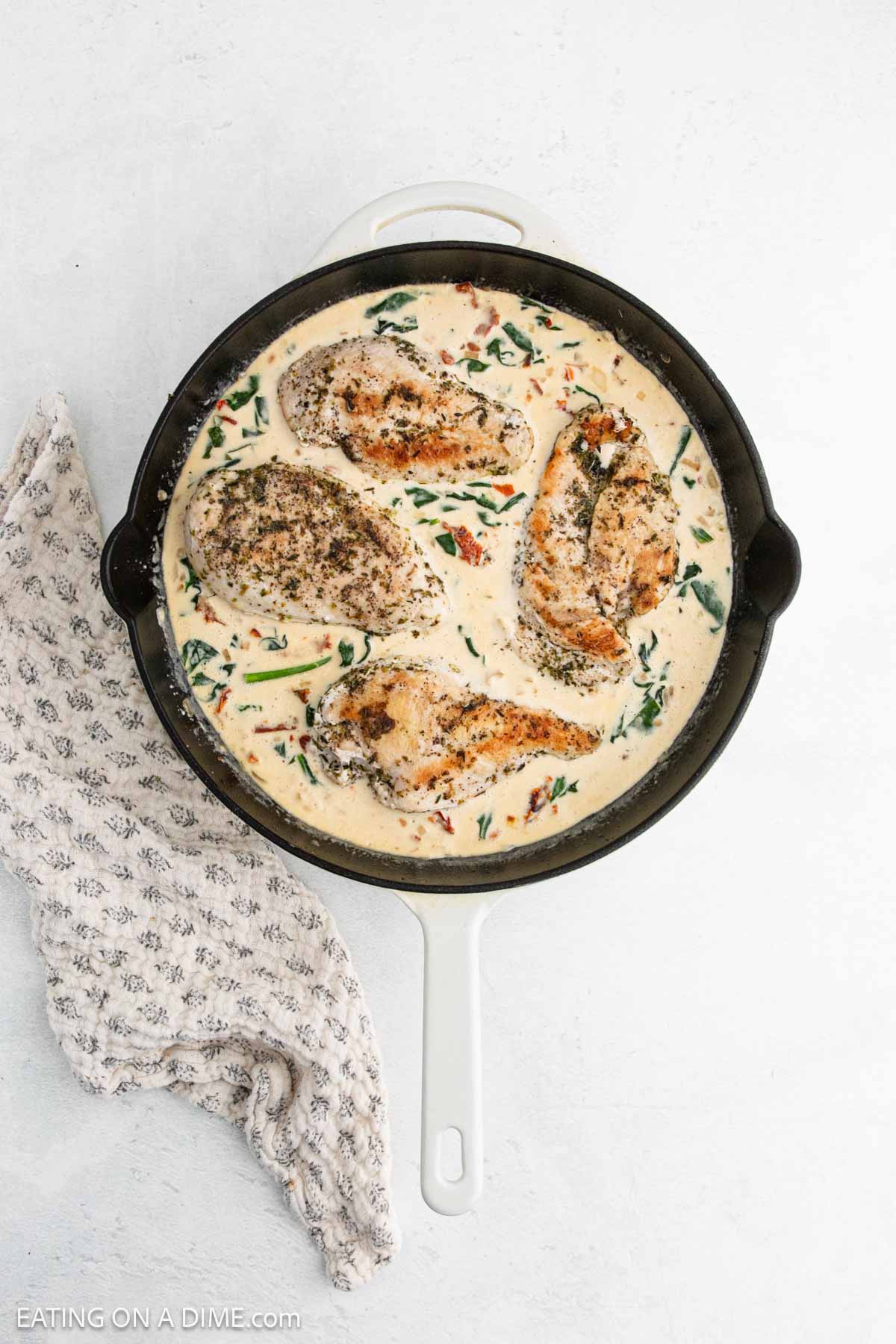 A skillet filled with creamy Tuscan Chicken, featuring seasoned chicken breasts in a rich sauce with spinach and sun-dried tomatoes. A patterned kitchen towel is beside the skillet on a white surface—an inviting Tuscan Chicken recipe.