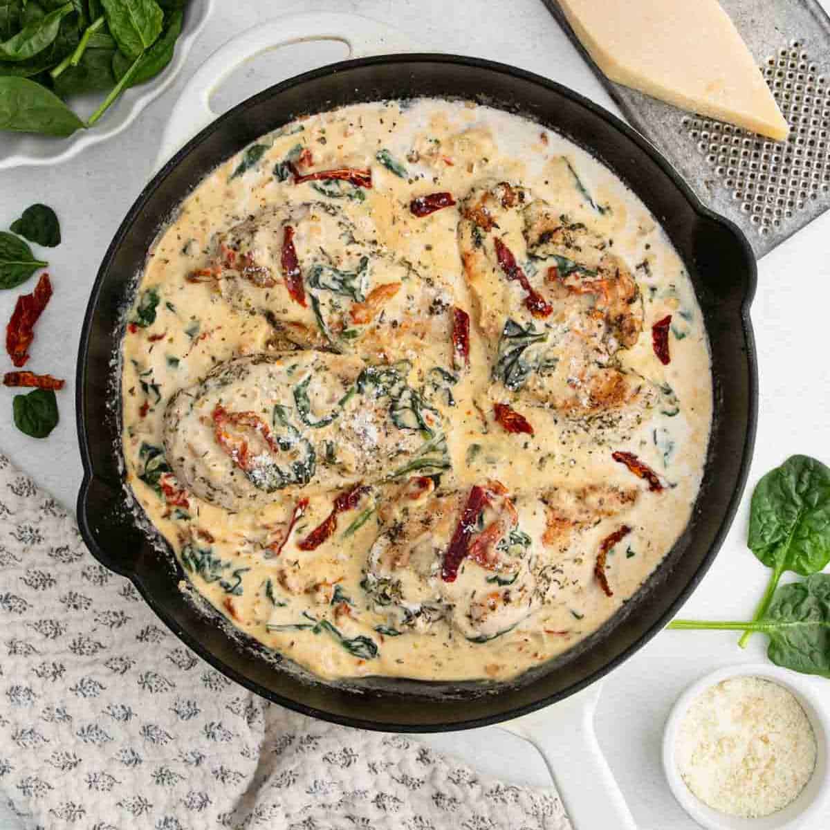 A skillet filled with creamy Tuscan Chicken, featuring seared chicken breasts in a rich cream sauce with spinach and sun-dried tomatoes. This Tuscan Chicken recipe is topped with grated cheese and fresh spinach on a white surface.