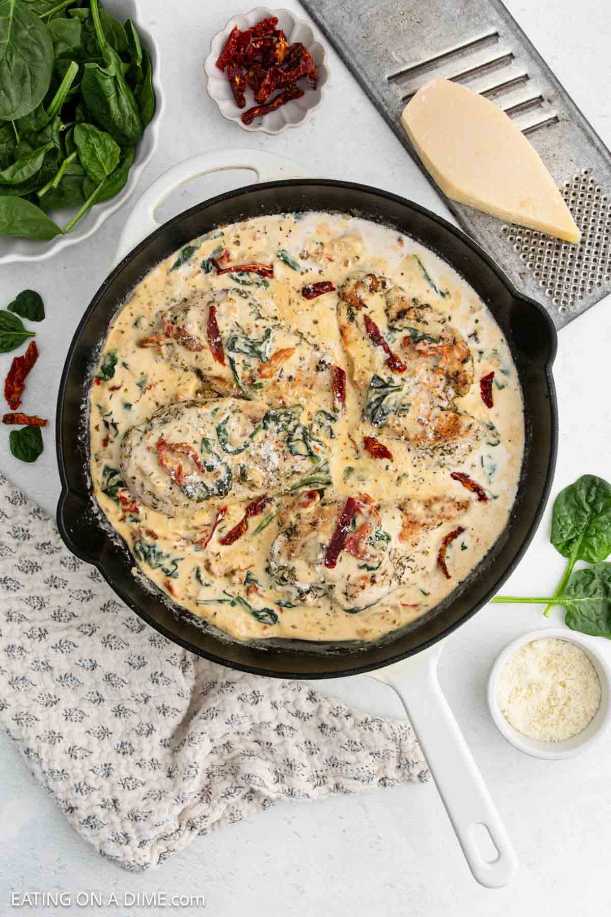 A skillet filled with creamy Tuscan Chicken simmers, featuring chicken breasts in a rich cream sauce with spinach and sun-dried tomatoes. This Tuscan Chicken Recipe shines with fresh spinach, grated cheese, and sun-dried tomatoes nearby on a white countertop.