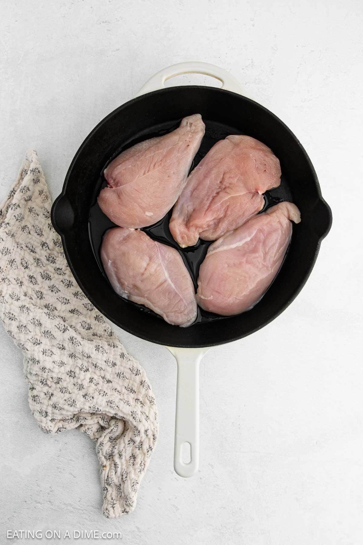 Four raw chicken breasts are arranged in a black cast iron skillet with a white handle, ready for your favorite Tuscan Chicken Recipe. A cream-colored patterned kitchen towel rests beside the skillet on a light gray surface.