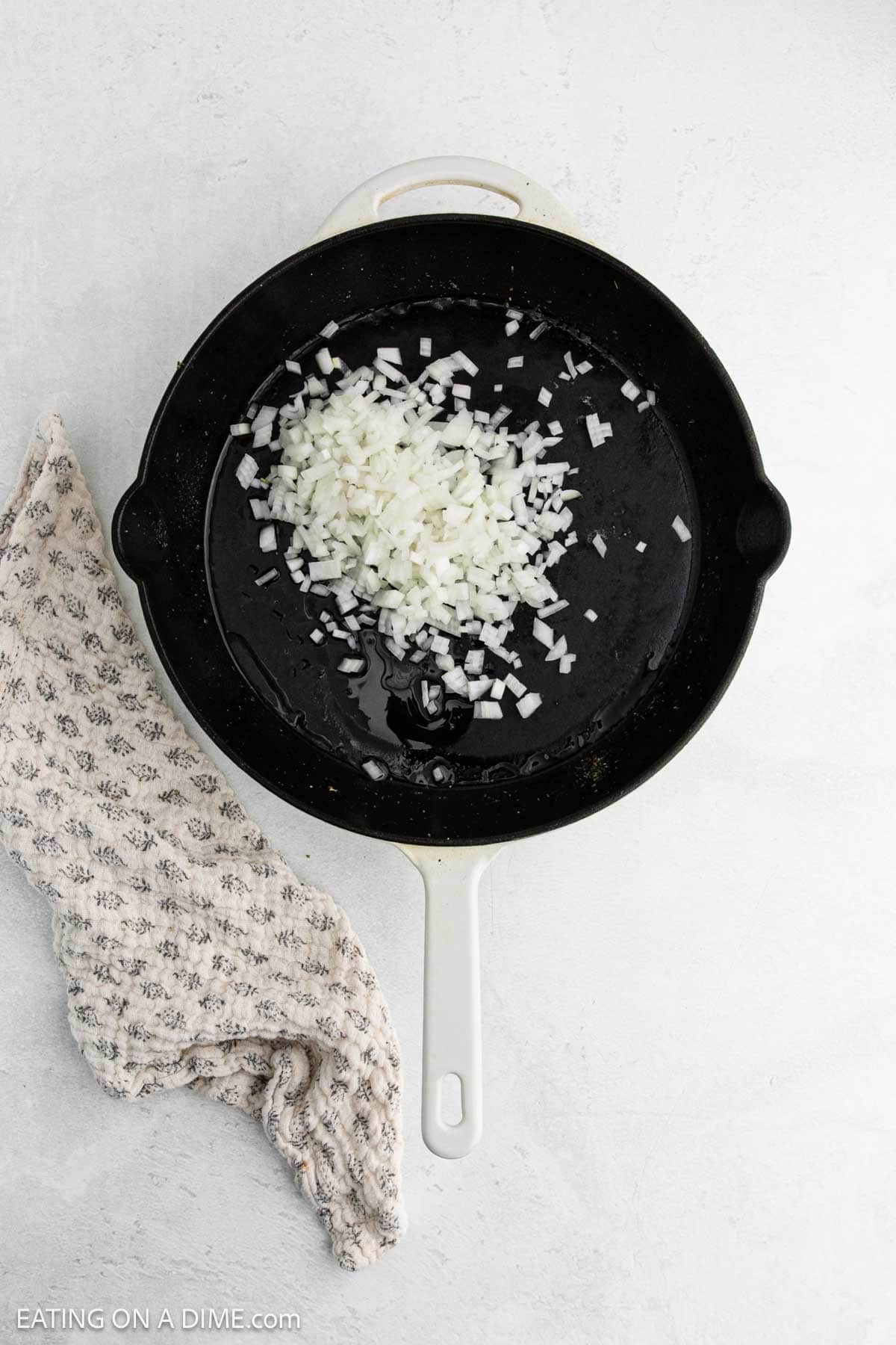 A white skillet with chopped onions and oil sits on a light surface next to a beige patterned oven mitt, ready for the first step in a delicious Tuscan chicken recipe.