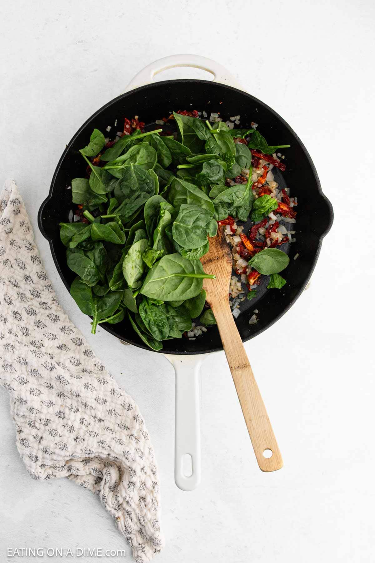 A white skillet filled with fresh spinach, diced red peppers, and onions sits on a white surface—essential ingredients for a flavorful Tuscan Chicken Recipe. A wooden spoon rests in the pan, with a textured cloth placed nearby.