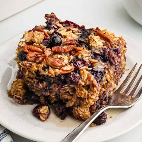 A slice of baked oatmeal, topped with juicy blueberries and crunchy pecans, sits on a white plate with a fork, highlighting its moist and hearty texture.