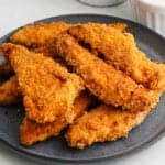 A dark plate holds a pile of golden-brown, crispy oven fried chicken tenders, with a small dish of dipping sauce visible in the background.