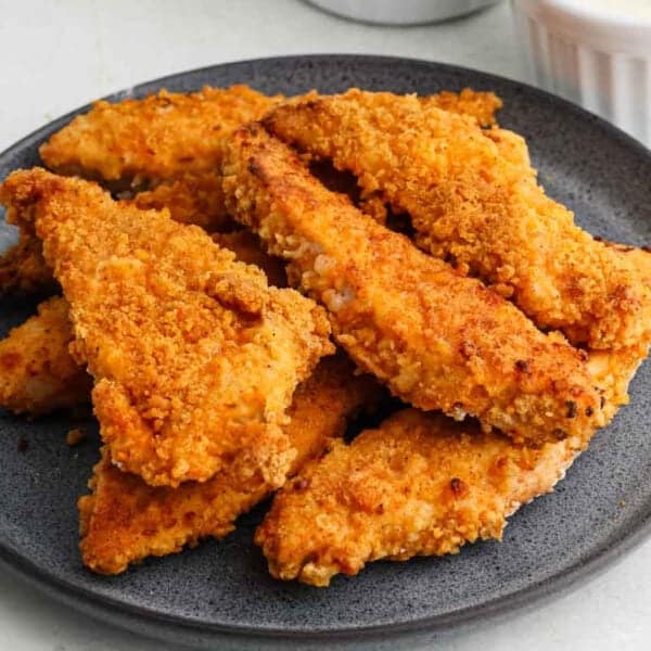 A dark plate holds a pile of golden-brown, crispy oven fried chicken tenders, with a small dish of dipping sauce visible in the background.