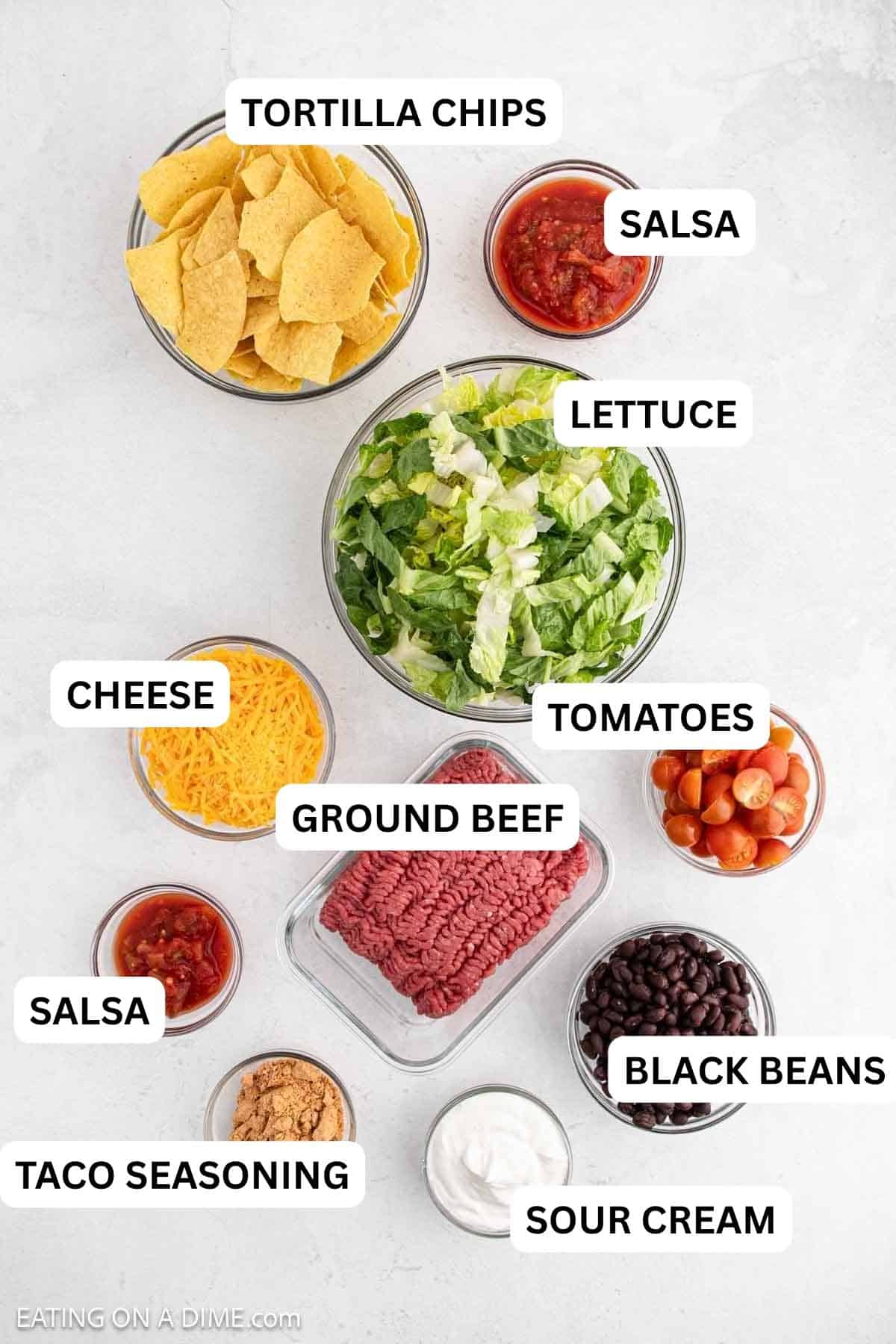 Ingredients arranged on a white background include tortilla chips, salsa, shredded cheddar cheese, ground meat, sour cream, black beans, cherry tomatoes, and a large bowl of chopped lettuce.
