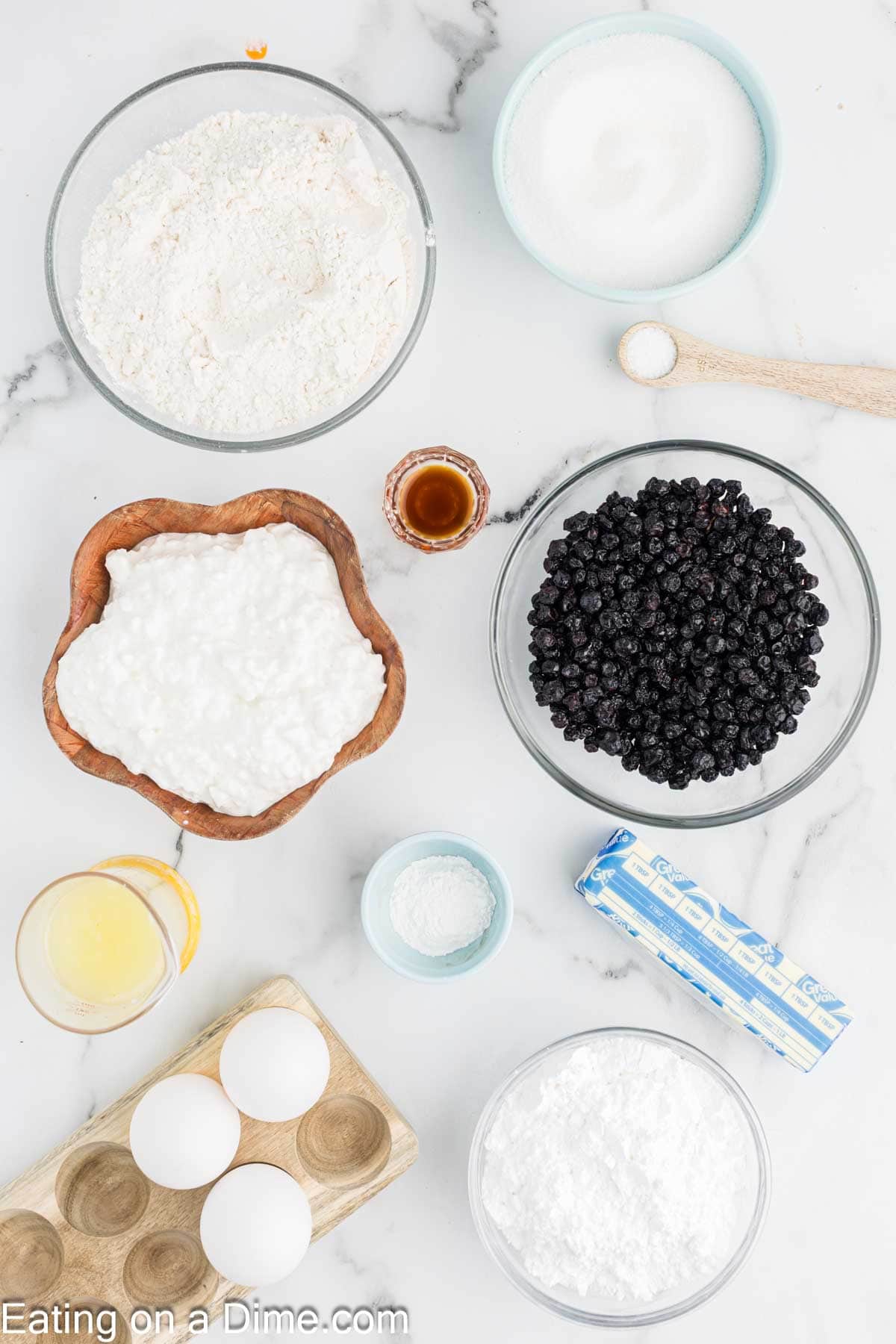 Top-down view of baking ingredients in bowls on a marble surface, perfect for making a Cottage Cheese Cake, including flour, sugar, eggs, butter, cottage cheese, vanilla, baking powder, lemon juice, powdered sugar, and fresh blueberries.