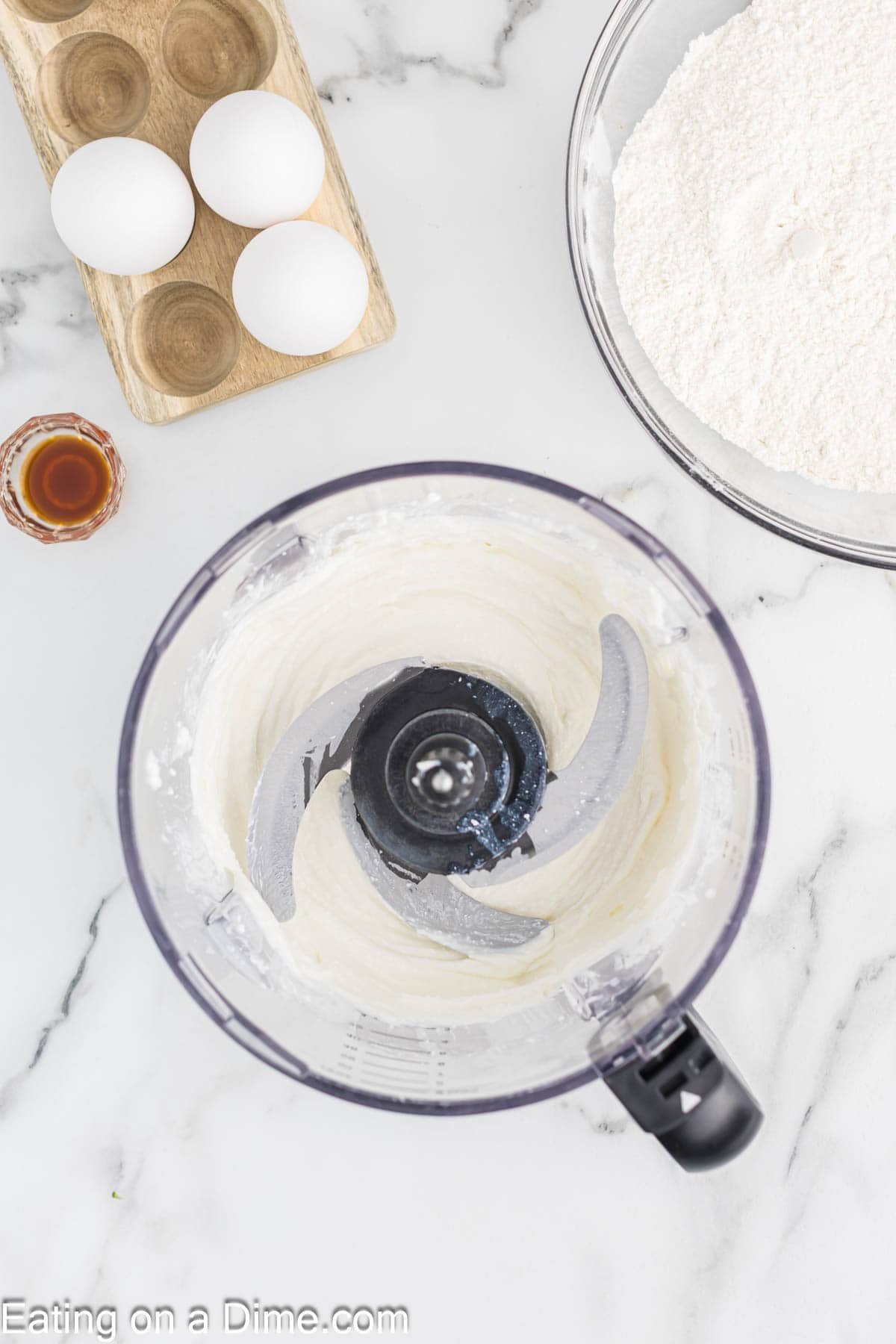 Overhead view of a food processor with creamy cottage cheese cake batter inside, next to a bowl of flour, a small bottle of vanilla extract, and a wooden tray holding three eggs on a marble countertop.