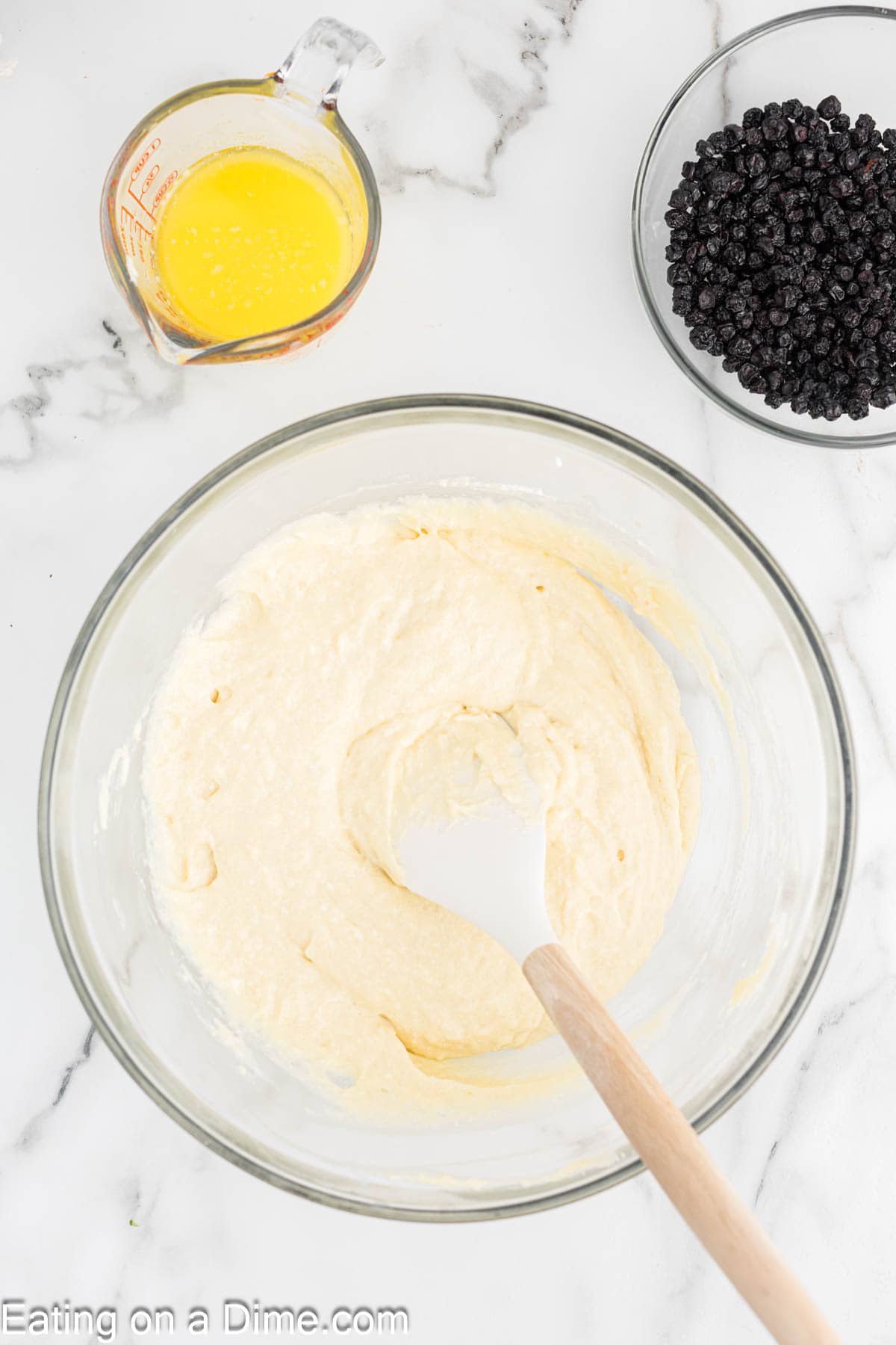 A glass bowl with muffin batter and a spatula, next to a small glass measuring cup of melted butter and a bowl of dried blueberries on a white marble surface—perfect for preparing Cottage Cheese Cake or blueberry muffins.