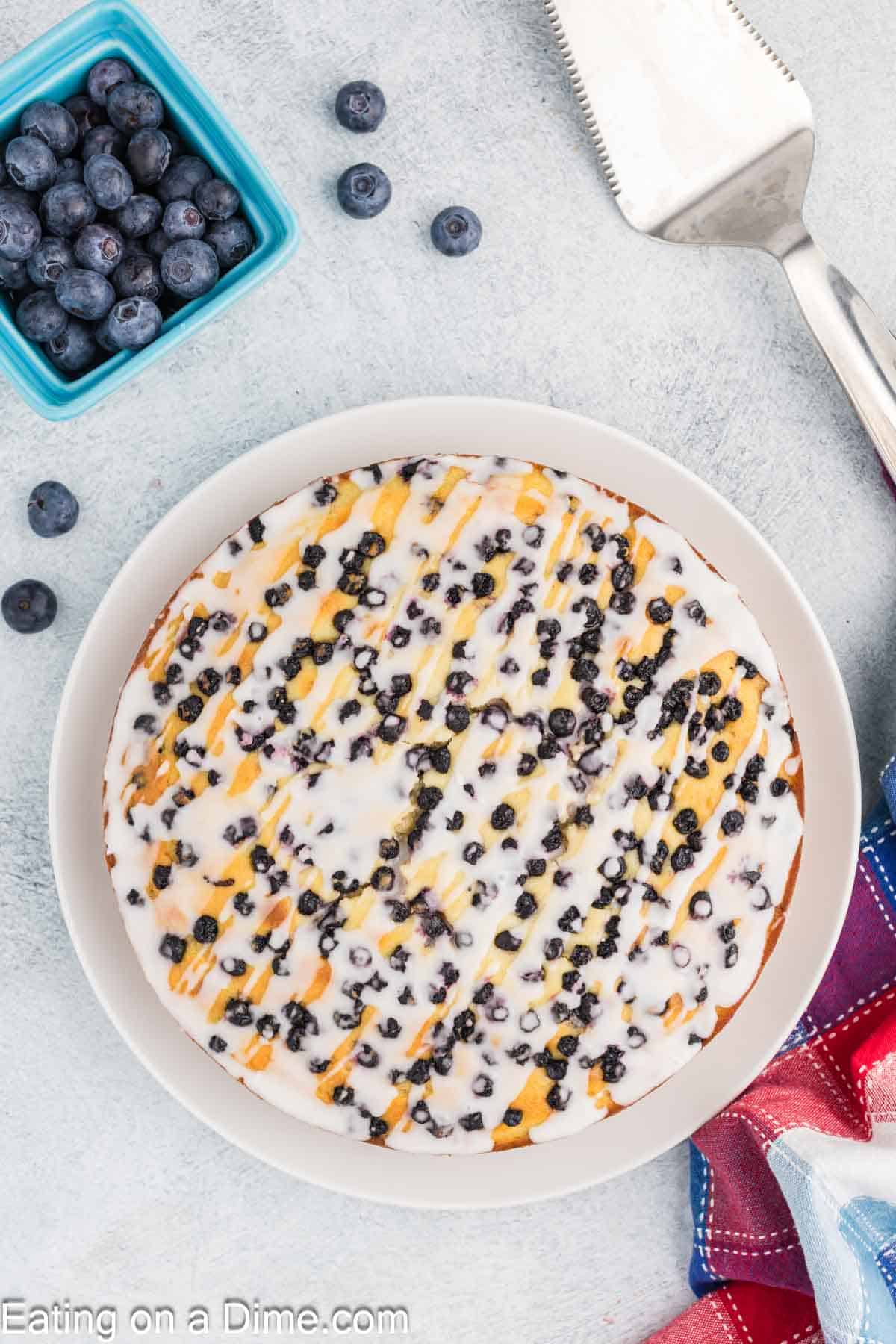 A round high protein blueberry cottage cheese cake with white icing drizzle sits on a white plate. Fresh blueberries are scattered on top, with more in a blue dish beside the cake. A metal cake server and a red-blue cloth are nearby.