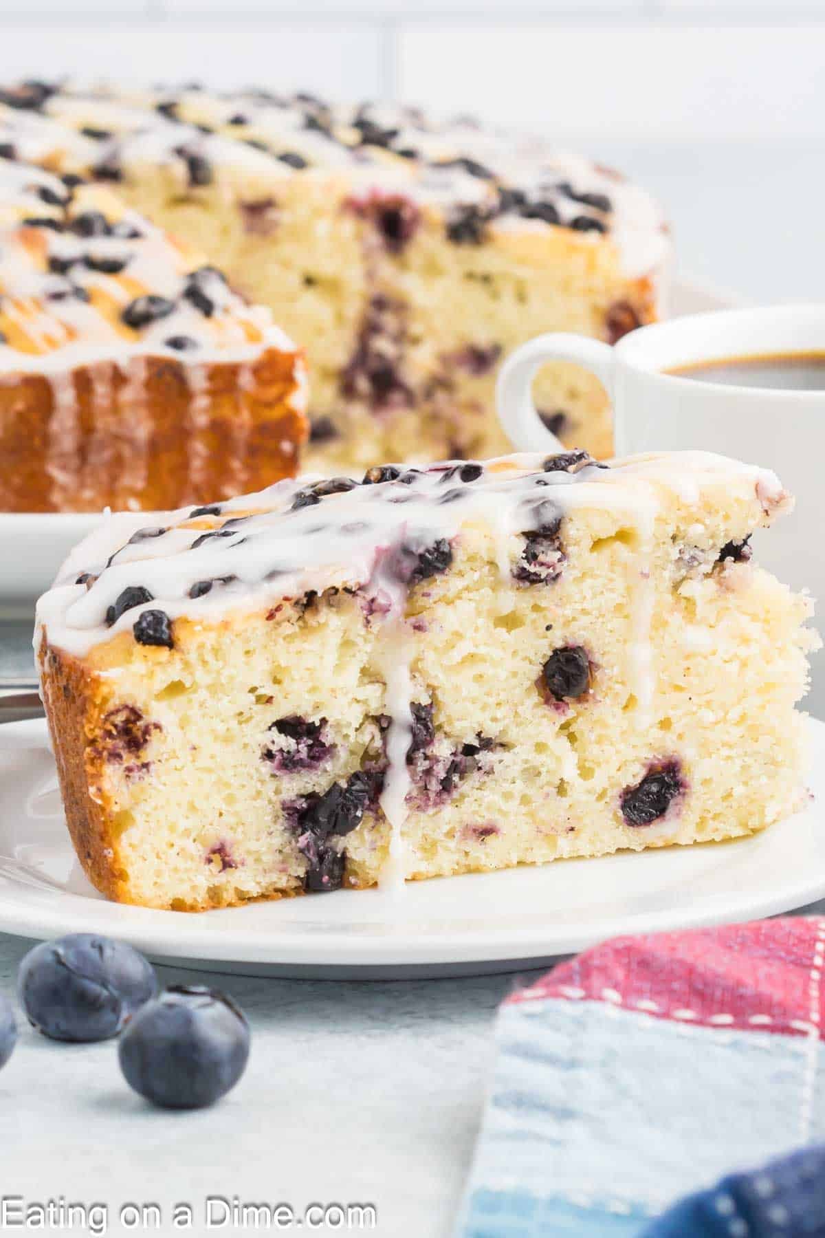A slice of high protein blueberry cottage cheese cake with white icing drizzled on top sits on a white plate next to a cup of coffee. Whole blueberries and a colorful napkin are nearby, with the rest of the cake in the background.