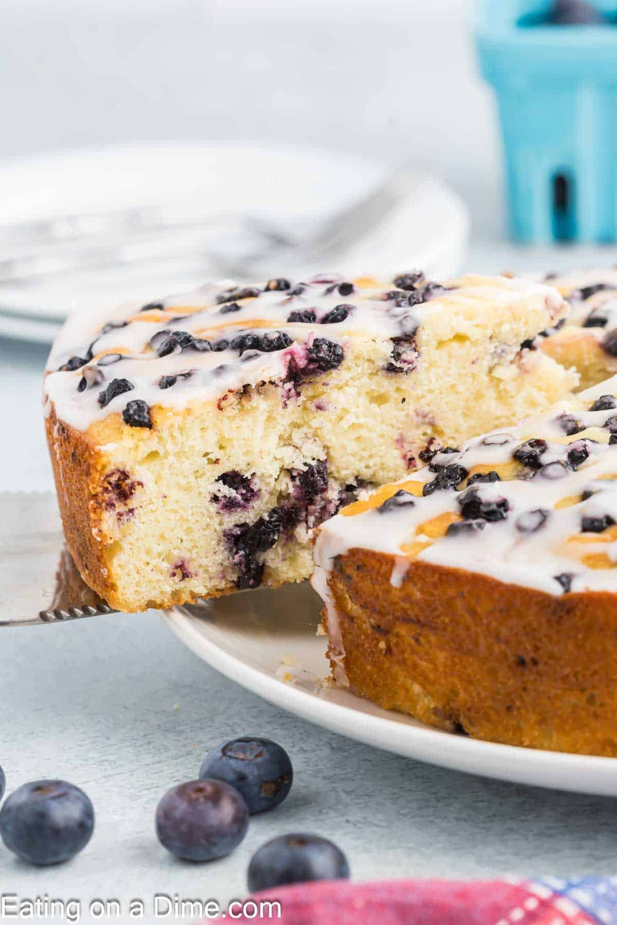 A slice of high protein blueberry cottage cheese cake with white icing is being lifted from a whole cake on a white plate. Fresh blueberries are scattered around, and a blue container of blueberries is in the background.