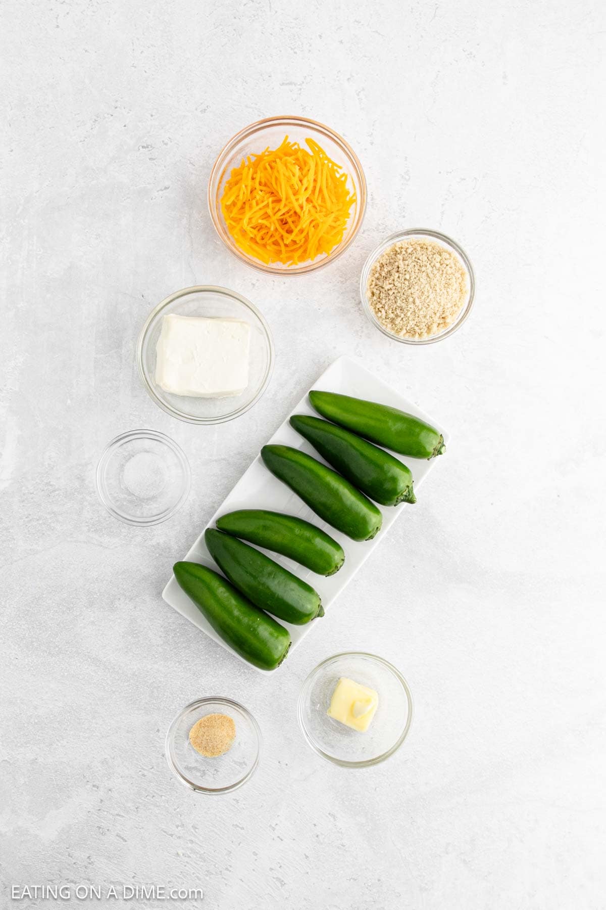 Six fresh jalapeños on a rectangular white plate are surrounded by small bowls of cheddar, cream cheese, breadcrumbs, butter, and seasoning—perfect for making delicious Air Fryer Jalapeño Poppers right at home.
