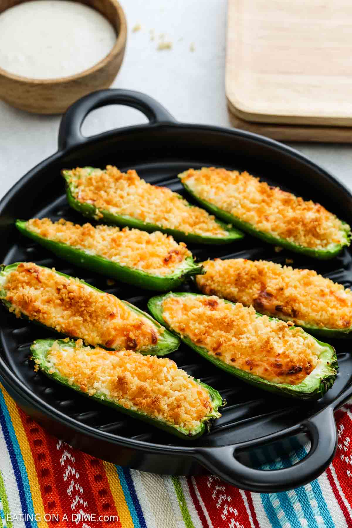 A round black plate holds several baked Jalapeño Poppers topped with golden brown breadcrumbs. A bowl of dipping sauce and a wooden cutting board are in the background, while the plate rests on a colorful woven cloth. Perfect for air fryer fans!.