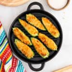 A round black grill pan holds seven Air Fryer Jalapeño Poppers with a golden, crispy topping. A colorful striped cloth and a bowl of white dipping sauce are nearby on a light surface.