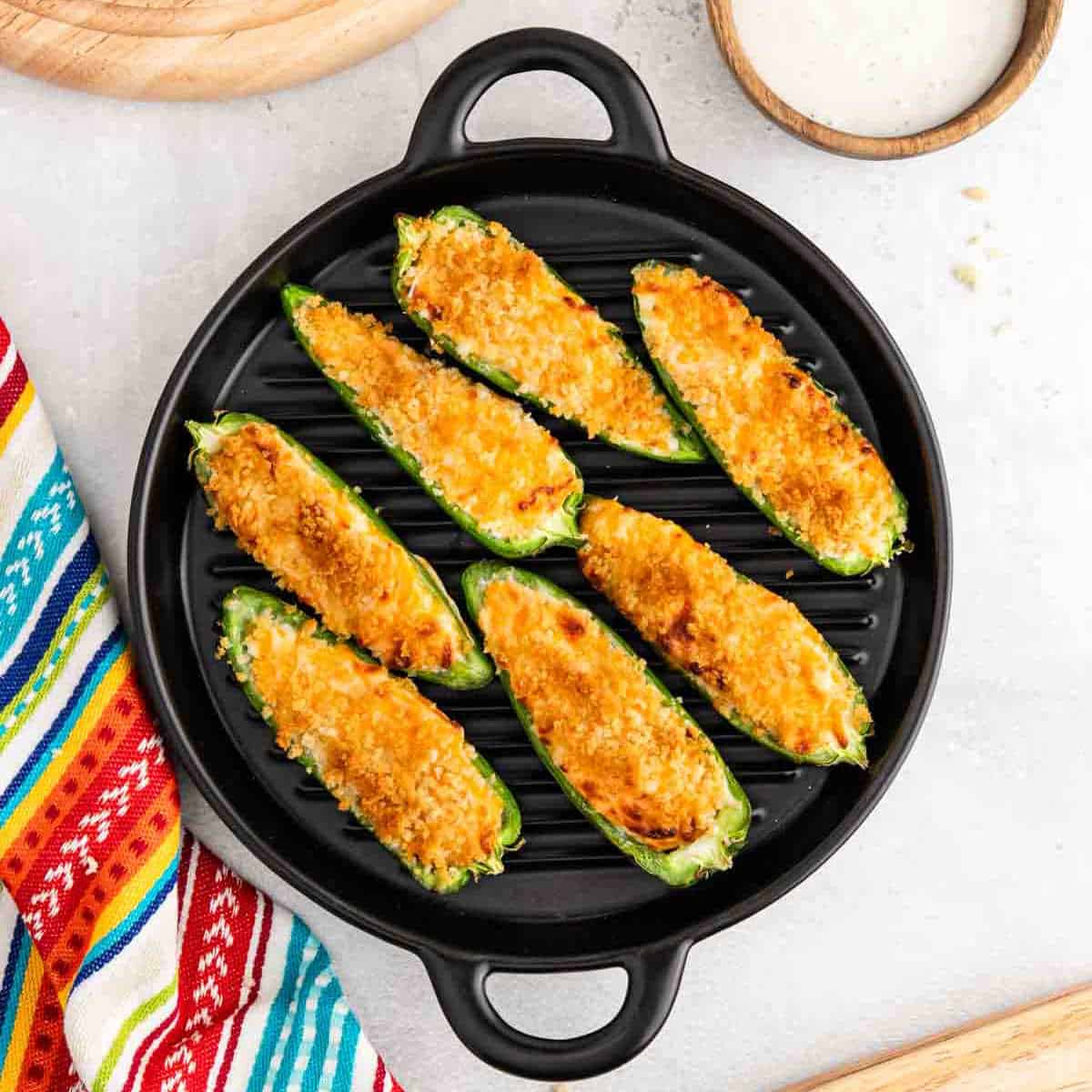A round black grill pan holds seven Air Fryer Jalapeño Poppers with a golden, crispy topping. A colorful striped cloth and a bowl of white dipping sauce are nearby on a light surface.