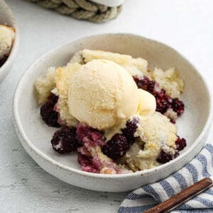 A bowl of Blackberry Cobbler topped with a scoop of vanilla ice cream, served on a white plate with a blue striped napkin nearby—a perfect blackberry dessert for any cobbler recipe lover.