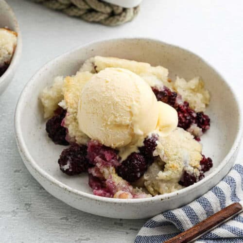 A bowl of Blackberry Cobbler topped with a scoop of vanilla ice cream, served on a white plate with a blue striped napkin nearby—a perfect blackberry dessert for any cobbler recipe lover.