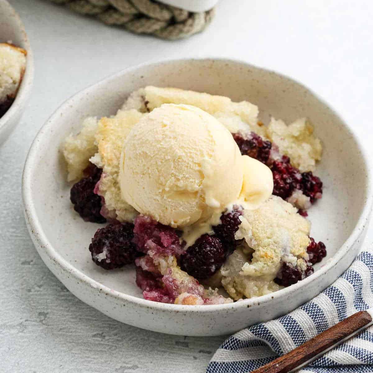 A bowl of Blackberry Cobbler topped with a scoop of vanilla ice cream, served on a white plate with a blue striped napkin nearby—a perfect blackberry dessert for any cobbler recipe lover.