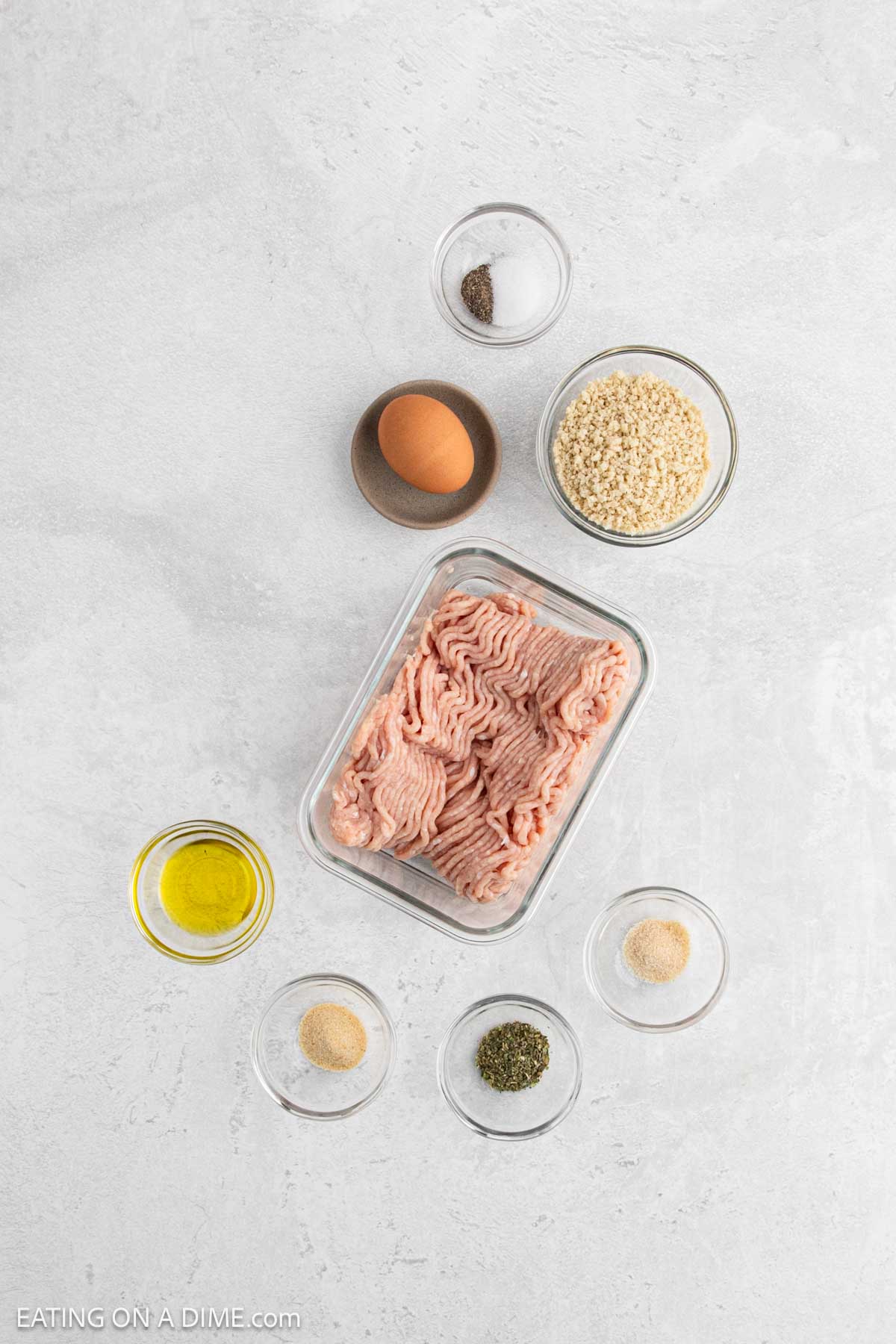 Overhead view of Chicken Meatballs ingredients, including ground turkey, an egg, dry breadcrumbs, oil, and various seasonings, each placed in small glass bowls on a light gray surface.