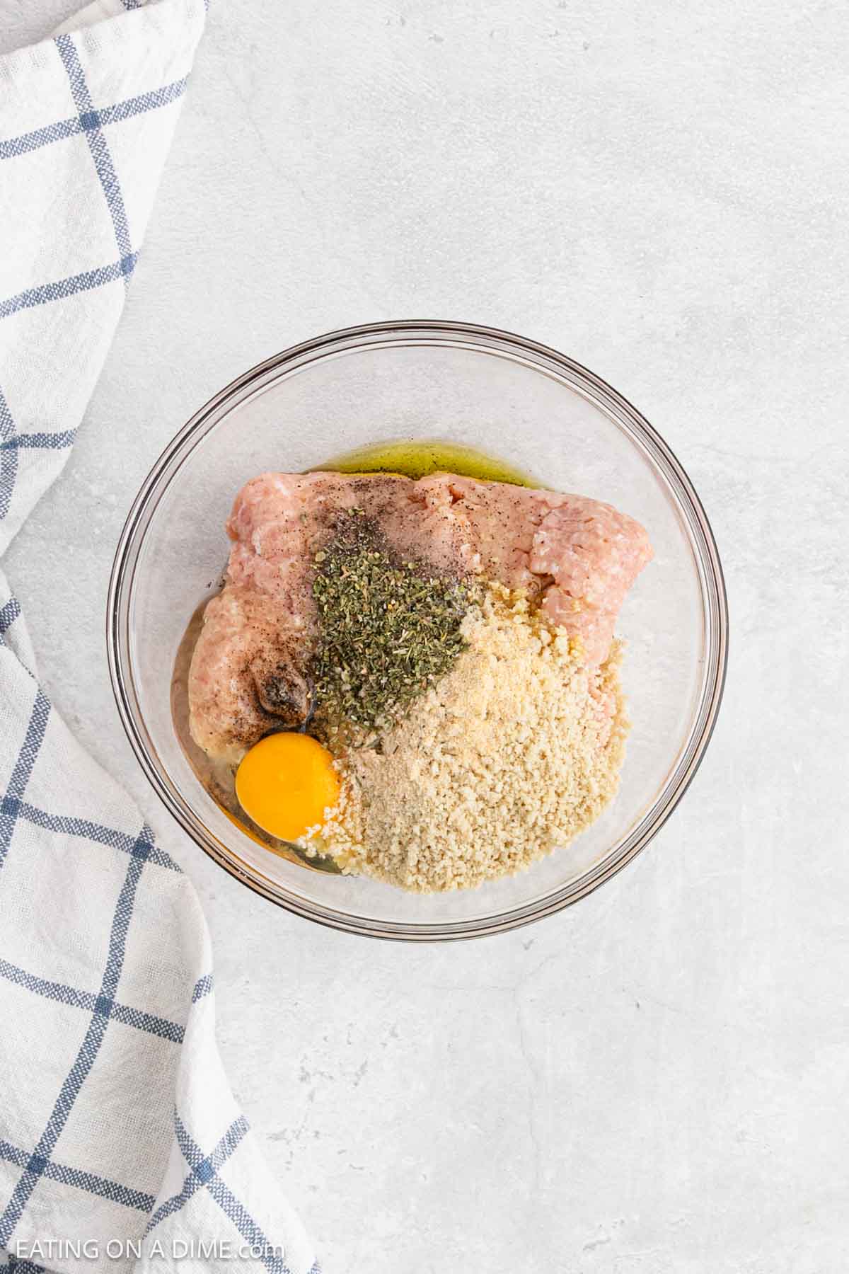 A glass bowl containing ground meat for a tasty meatball recipe, a cracked egg, breadcrumbs, dried herbs, and spices sits on a light countertop next to a white and blue checkered kitchen towel.