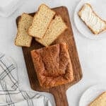 A loaf of Cottage Cheese Bread with a golden-brown crust sits on a wooden cutting board, several slices cut. A plate nearby holds a buttered slice, while a checkered cloth is in the corner.