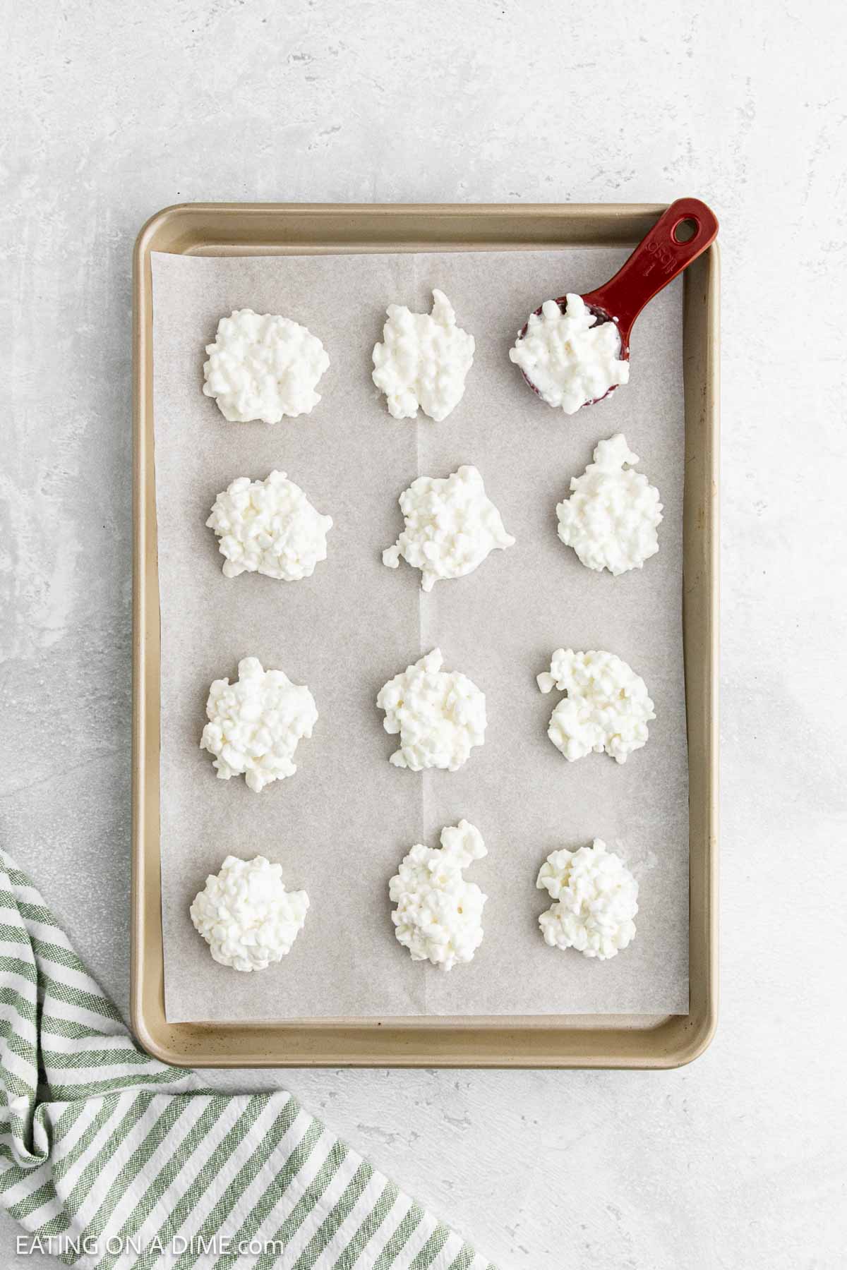 A baking sheet lined with parchment paper holds 12 clusters of a white, textured mixture resembling Cottage Cheese Chips. A red spoon rests on the sheet, and a green-striped towel is nearby.