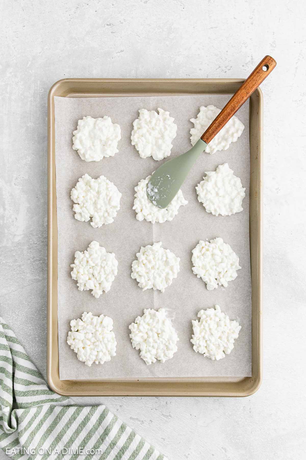 A baking sheet lined with parchment paper holds twelve clusters of white no-bake cookies, resembling Cottage Cheese Chips. A spatula with a wooden handle rests on the sheet, and a green and white striped towel is in the corner.