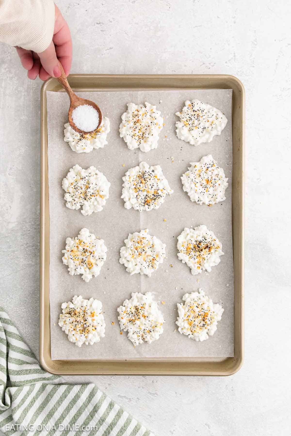A hand sprinkles salt over clusters of cottage cheese chips on a parchment-lined baking sheet. The chips are topped with seasoning and neatly arranged in rows, while a striped towel is partially visible in the corner.