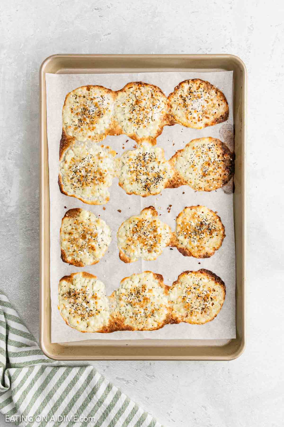 A baking sheet with parchment paper holds several golden-brown, round cottage cheese chips sprinkled with seasoning. A green and white striped towel is visible in the bottom left corner.